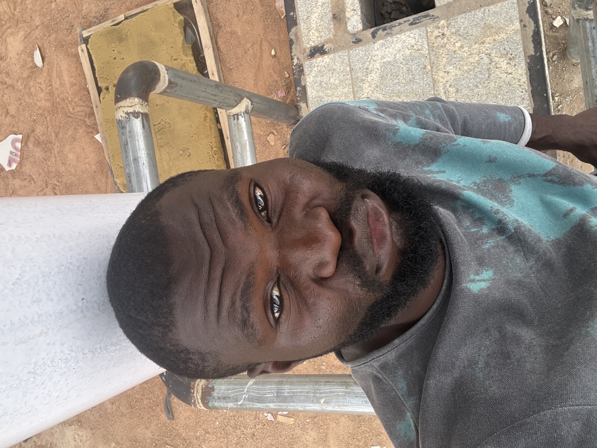 A close-up shot features an adult male with dark skin, a closely-cropped hairstyle, and a beard. He is wearing a grey and teal tie-dye shirt. The background includes a metal pipe structure, a yellow rectangular object within a wooden frame, tiled ground, and soil. It appears to be an outdoor setting. The location is specified as Barakin Lerawi, Nigeria. No action or interaction can be identified.
