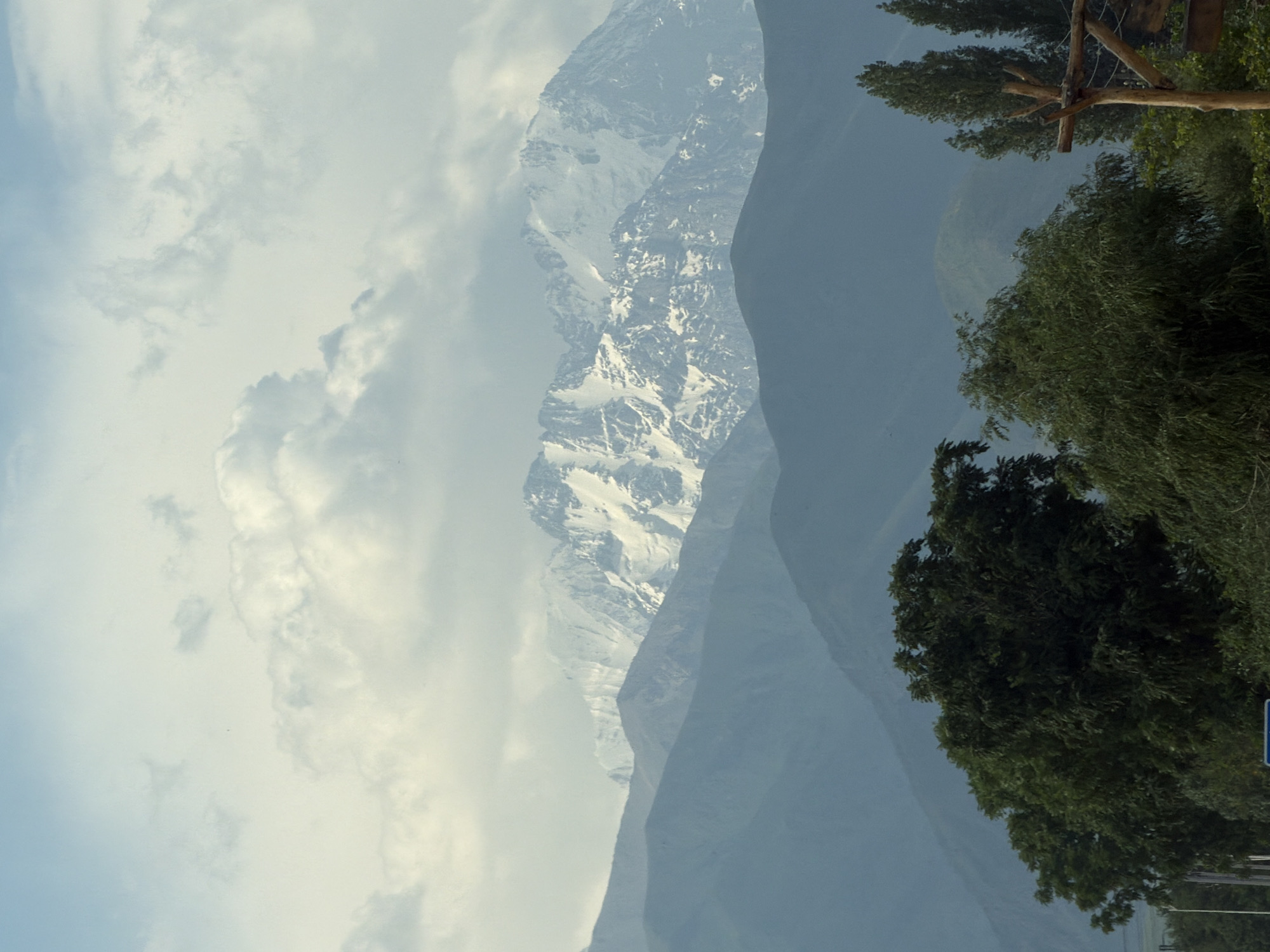 The image depicts a landscape scene with multiple layers of mountains stretching into the distance. The foremost layer consists of dark green, dense trees, with some lighter green foliage visible. Beyond these trees, a series of progressively lighter blue-toned mountains form layered silhouettes. The background is dominated by a prominent mountain range covered in snow, with rugged, rocky terrain visible through patches of snow. Above the mountains, the sky is filled with a mix of light blue and soft, diffused white clouds, suggesting an overcast or partly cloudy day. The lighting is soft and diffused, with no harsh shadows, indicating that the sun may be obscured by clouds. The overall impression is one of a vast, natural landscape with a sense of depth and scale. There are no people, animals, or man-made structures readily apparent in the main body of the image.