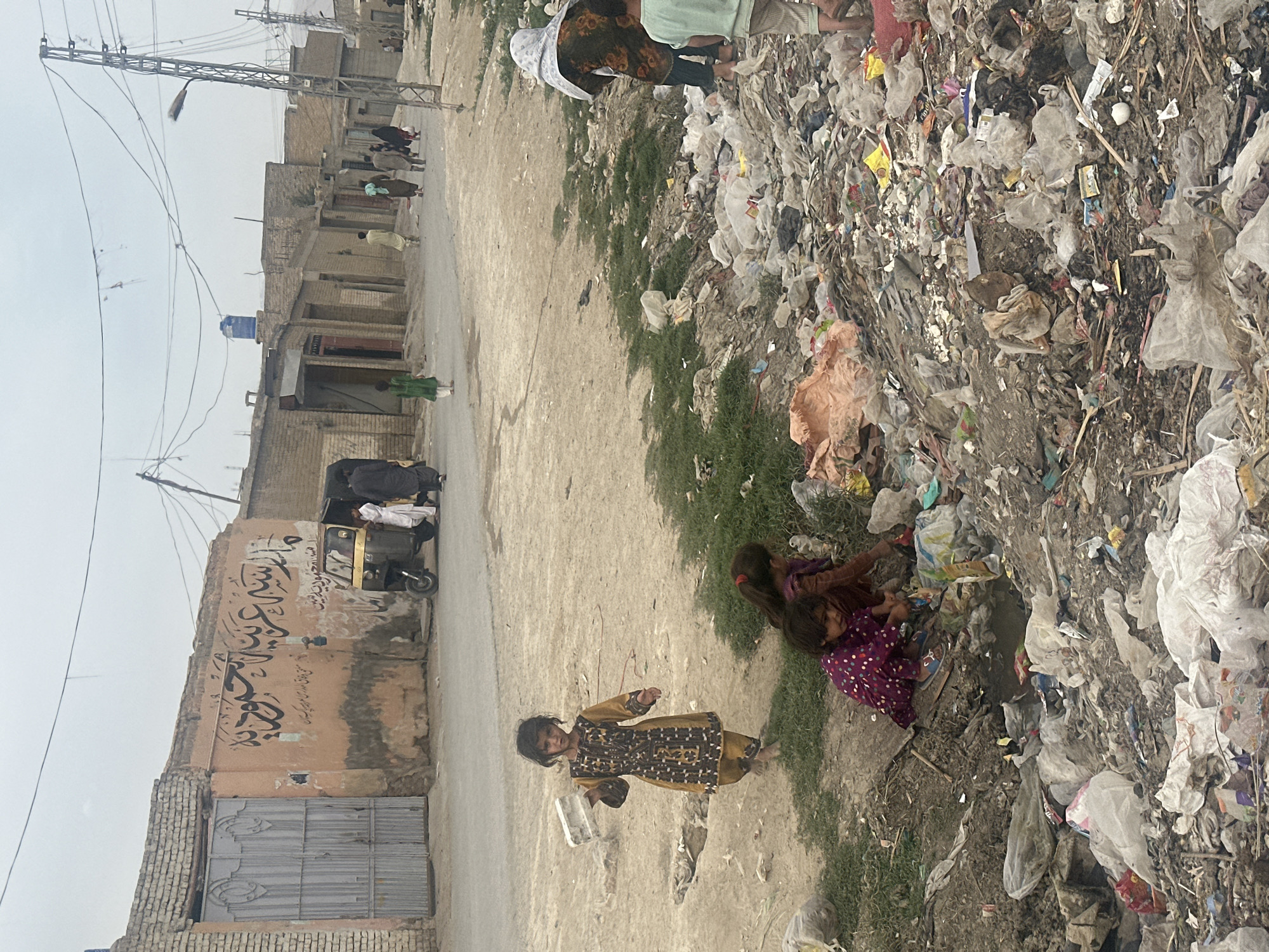 The scene is a street in Quetta City Tehsil, Pakistan. The street is lined with buildings of brick and concrete. One building has painted calligraphy. An auto rickshaw vehicle is parked nearby. Adjacent to the street is a large pile of garbage made up of plastic bags and general refuse. Several children are near the garbage. One is holding a plastic container. Two other children are sitting. Other people are walking in the distance. The environment is overcast.