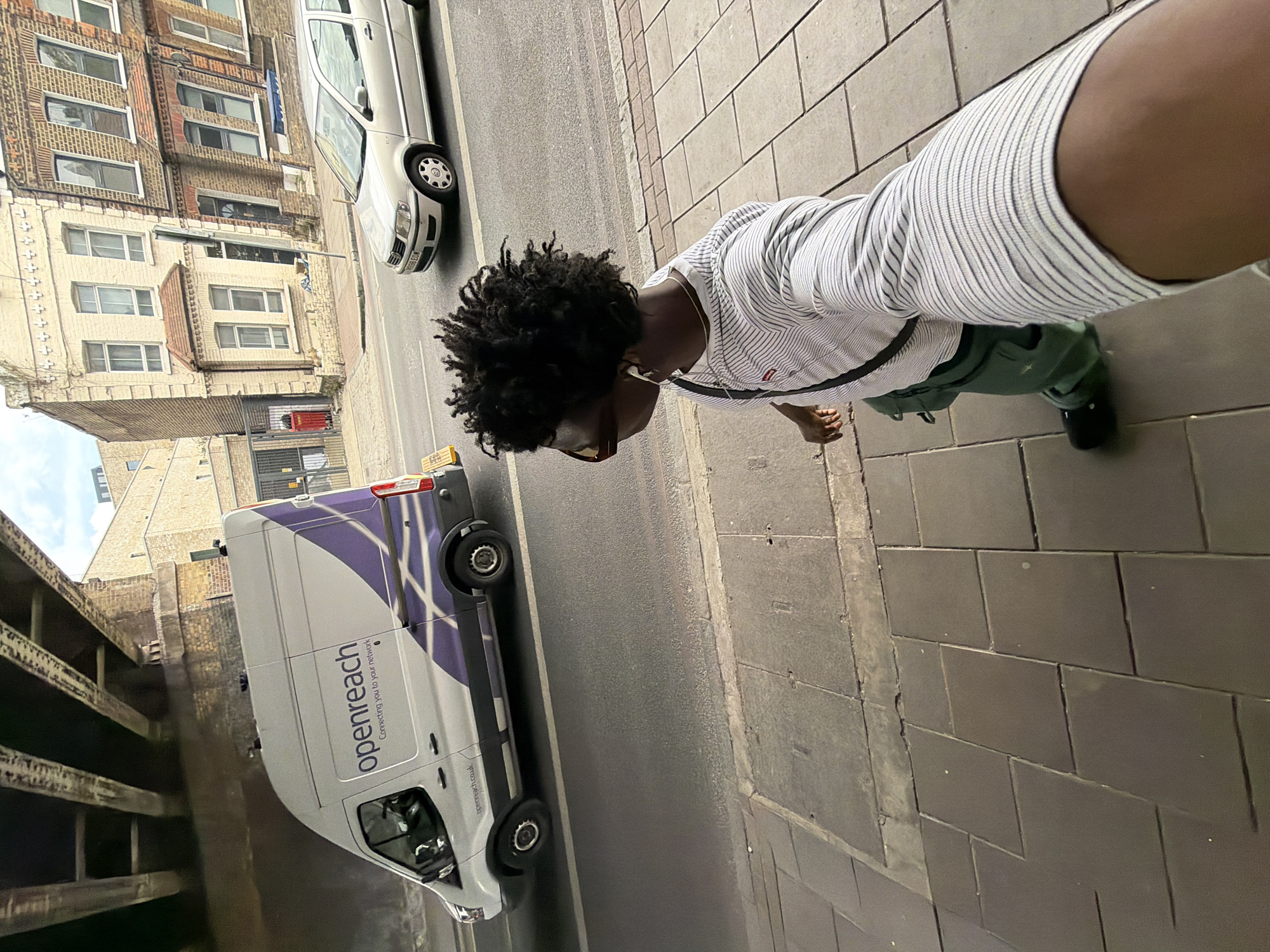 A medium shot in an urban area of London, United Kingdom. A person with dark, curly hair is positioned on a paved sidewalk, holding a camera phone up to take a selfie. The person wears sunglasses, a striped shirt, green pants, and black shoes. A silver Volkswagen car and a white Openreach van with purple stripes are parked on the adjacent street. Buildings with brick facades and numerous windows line the opposite side of the street. A gray concrete overpass is visible on the left side of the frame.