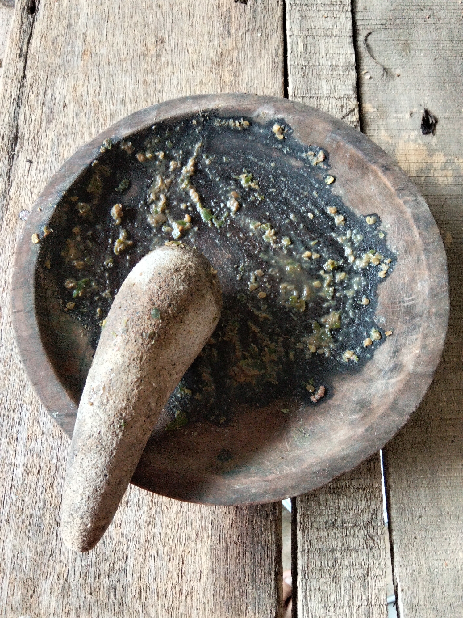 The image shows a rustic wooden mortar and pestle on a weathered wooden surface. The mortar is round and appears to be made of dark wood, with remnants of a ground mixture of spices or herbs visible inside. The pestle, made of a lighter, rougher material, is positioned diagonally across the mortar. The wooden surface beneath is composed of planks with visible grain and some imperfections, suggesting an outdoor or workshop setting. The scene is well-lit, with natural light casting shadows and highlighting the textures of the materials. The image does not contain any people or specific indications of a city location, therefore, its location in Benuaraja, Indonesia cannot be confirmed by the visual evidence alone.