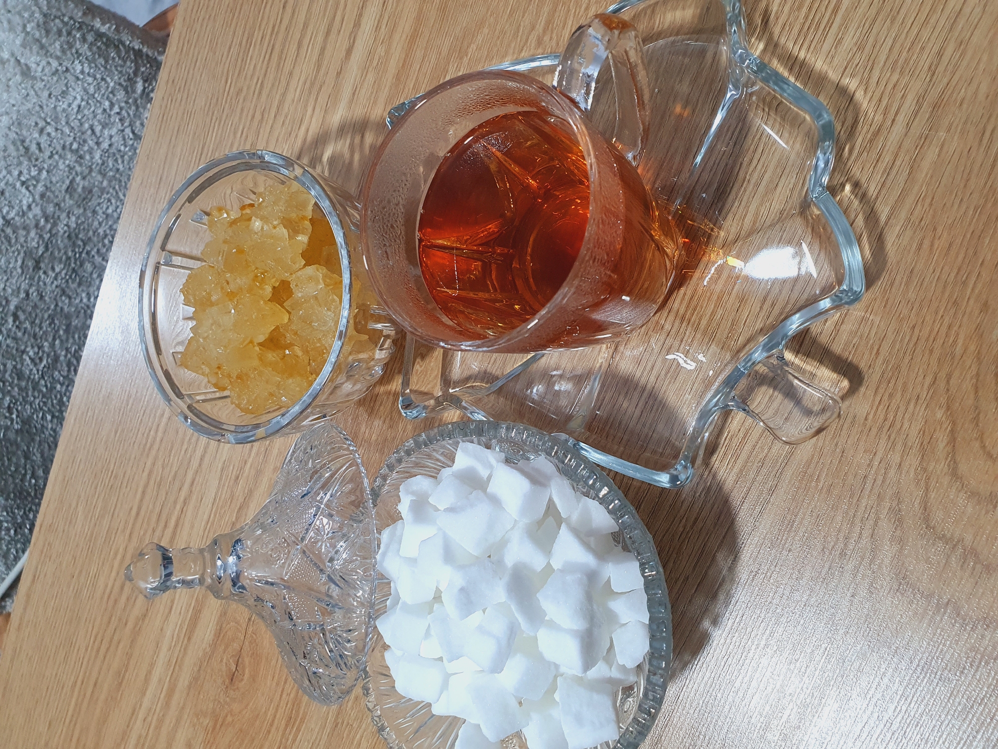 A glass of amber-colored tea sits on a clear glass saucer with a leaf-like design. Next to the tea, a clear glass bowl contains white sugar cubes, and another clear glass bowl holds amber-colored rock candy. A decorative crystal lid with a knob sits beside the bowl of sugar cubes. The objects are arranged on a light brown wooden surface. The background consists of a grey, textured fabric. The image is cropped from a wider view, with the top right corner showing a portion of the grey fabric. The scene appears to be indoors, possibly on a table. The time of day is not discernible. There are no people visible in the image. The location is identified as Kheyrabad, Iran.