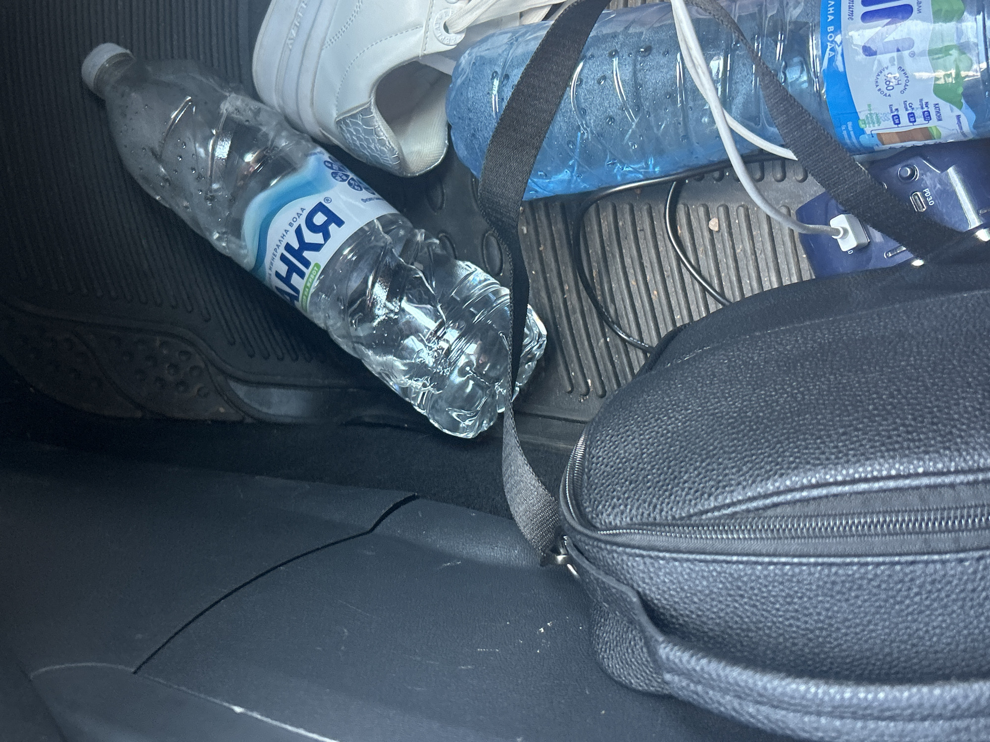 The image shows the interior of a vehicle, likely the rear seat or trunk area, in Plovdiv, Bulgaria. A black leather-textured bag with a zipper is in the foreground on the left. To the right of the bag are two plastic bottles of water, one larger and partially filled, the other smaller and wrapped in blue plastic. White sneakers are visible next to the larger water bottle. A portable power bank, connected by cables to other devices (not fully visible), is situated between the bag and the bottles. The floor of the vehicle is covered with a black rubber mat. The overall scene is cluttered, suggesting personal belongings stored within the car.