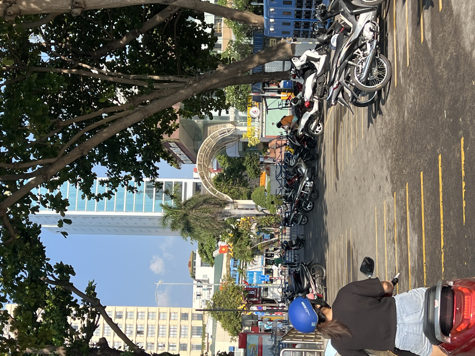The image depicts a street scene in Nha Trang, Vietnam. A person wearing a blue helmet and a dark-colored sweater is sitting on a red motorbike, facing away from the camera. The motorbike is positioned in a parking area lined with rows of parked motorbikes. The parking area is marked with yellow lines. In the background, there is a view of a city street with buildings, palm trees, and an arched structure. A Vietnamese flag is visible among the buildings.