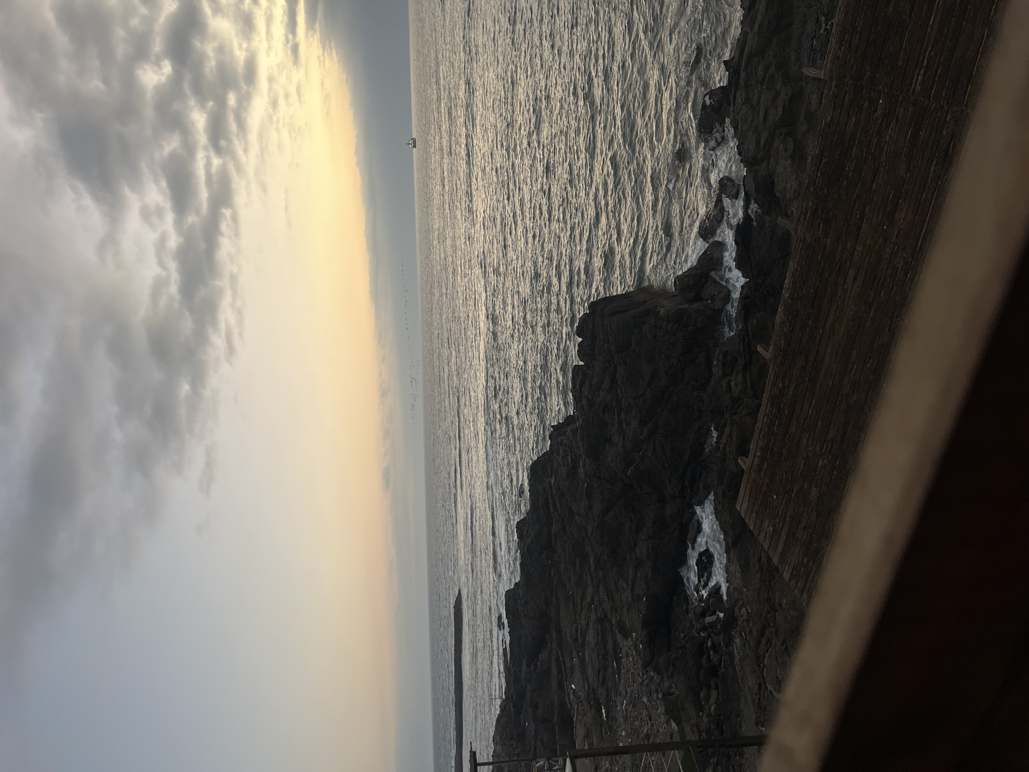 This is a landscape photograph taken at dusk or dawn. The foreground is dominated by dark, jagged rocks along the coastline and a wooden structure, possibly part of a building or a fence. In the distance is the ocean, with sunlight reflecting off the waves. A flock of birds is visible, and a small ship or boat in the distance. The sky is a blend of grays and yellows, suggesting either a sunrise or sunset. The location is likely along the coast in Arica, Chile.