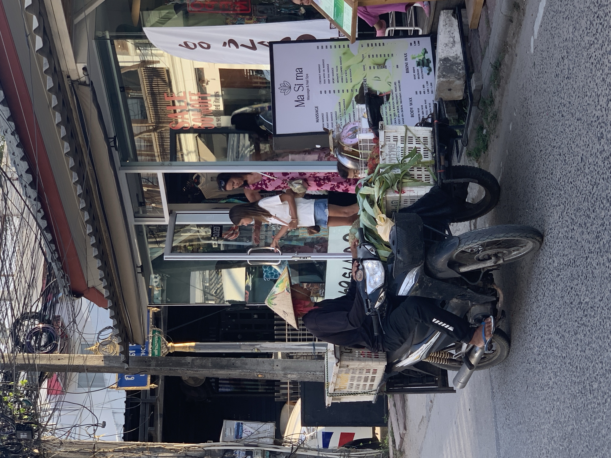 In Ko Samui District, Thailand, a person wearing a conical hat and black clothing is riding a three-wheeled motorcycle with a cargo rack in the front and back. The cargo rack in the front carries white plastic baskets filled with green produce, possibly vegetables or fruit. The motorcycle is black and appears to be modified with a larger cargo area. The person's hair is bright red and they are wearing a conical hat that appears to be hand-painted. The motorcycle is parked on a paved road in front of a storefront.
The storefront is a business with a glass door and windows. A "CLOSED" sign is visible on the door. Inside the store, several people are visible, including two women and a child. One woman is wearing a pink dress, and the other is wearing a white shirt and blue skirt. They appear to be interacting with each other. A sign with the text "SALE 50% OFF on selected items" is visible on the window. Next to the entrance, a large white sign displays prices for various services, including "Ma Si ma Massage & Nail Spa," and lists prices for "MASSAGE," "PEDICURE," "BODY WAX," and "BIKINI WAX."
The environment is characteristic of a bustling street scene with overhead electrical wires crisscrossing the sky. The buildings have awnings that provide shade. The road is asphalt, and there are some scattered items on the side of the road, including a concrete block. The lighting suggests it is daytime, possibly late afternoon due to the shadows.