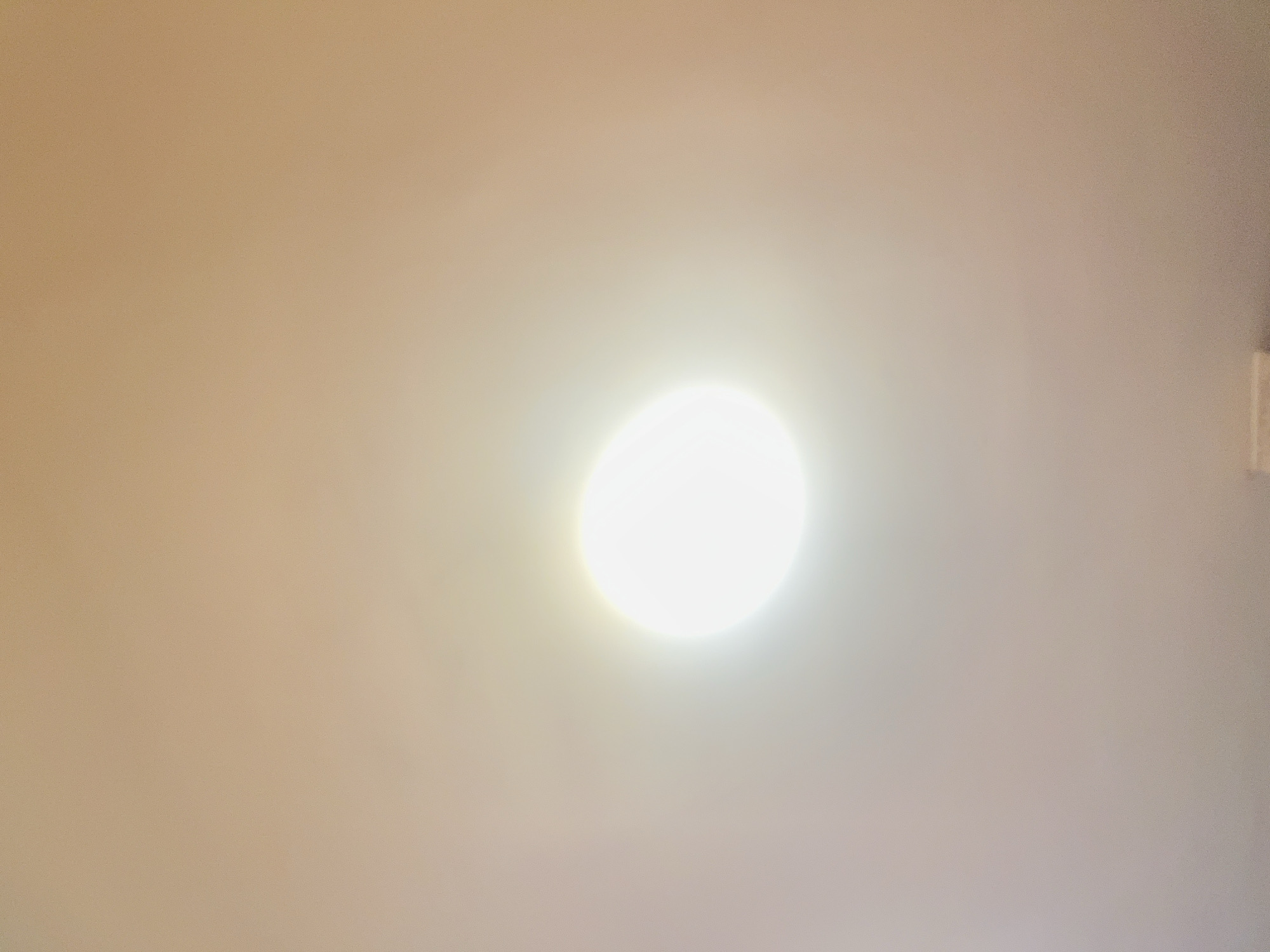 The image shows a bright, circular light source against a beige/tan background. The light source is positioned near the center of the frame, with the background appearing slightly darker towards the edges. A small, rectangular object is visible in the upper right corner, near the edge of the frame. There are no people, subjects, objects, actions, or interactions visible in the image, other than the light source and the background. The location is unknown and impossible to determine from this image.