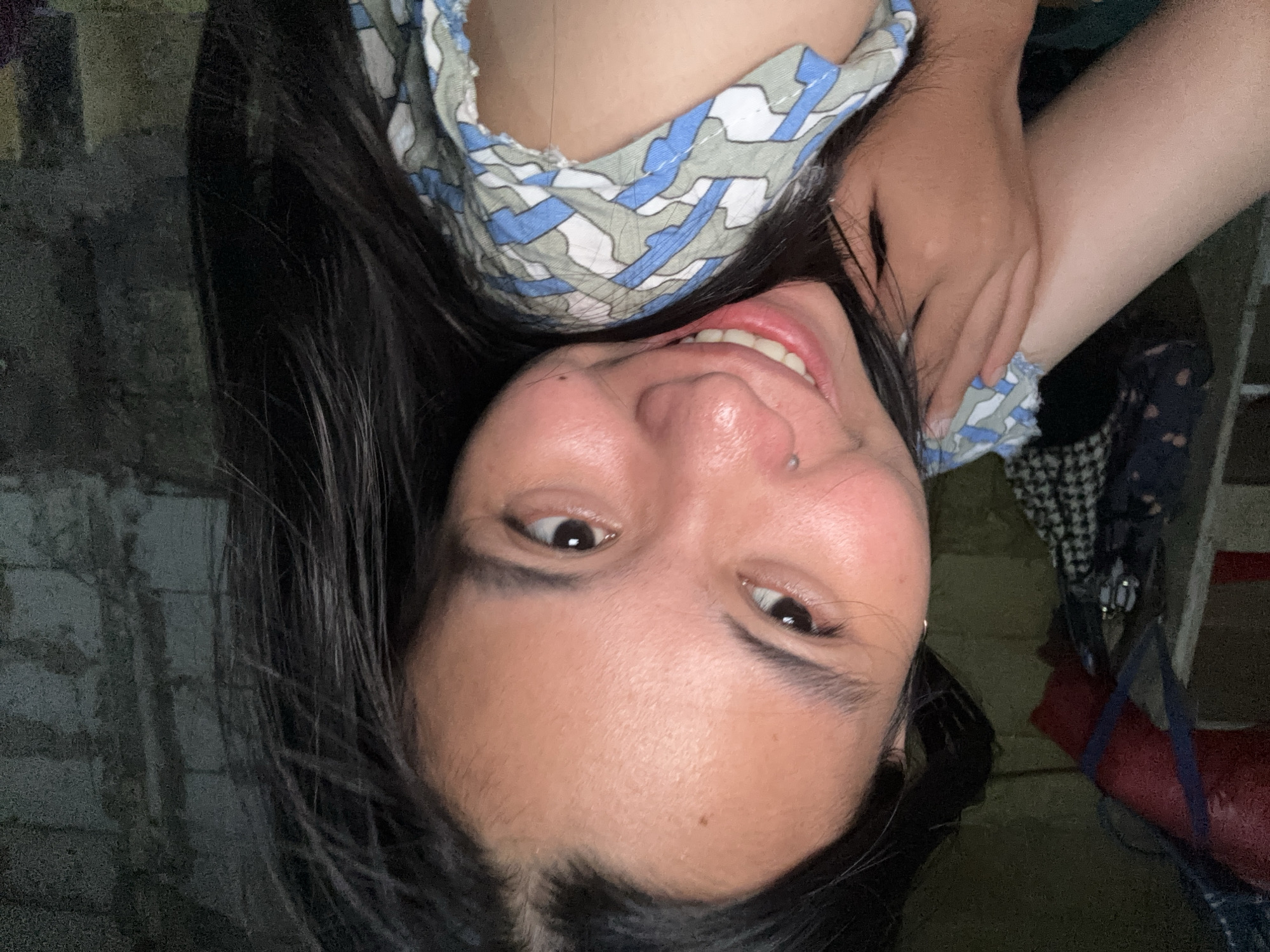 This is a close-up, eye-level, medium shot of a young woman with long, dark hair smiling directly at the camera. She is wearing a light-colored shirt with a blue, white, and grey abstract pattern. Her left arm is bent and resting across her chest, with her hand near her neck. She has a small silver stud piercing on her nose and a subtle mole on her cheek. The background is dark and out of focus, showing what appears to be a rough, textured wall and some hanging items. The lighting is dim, creating a soft glow on the woman's face. The overall impression is a casual, self-taken photograph. There is no discernible information in the image to suggest a location in East Jakarta, Indonesia.