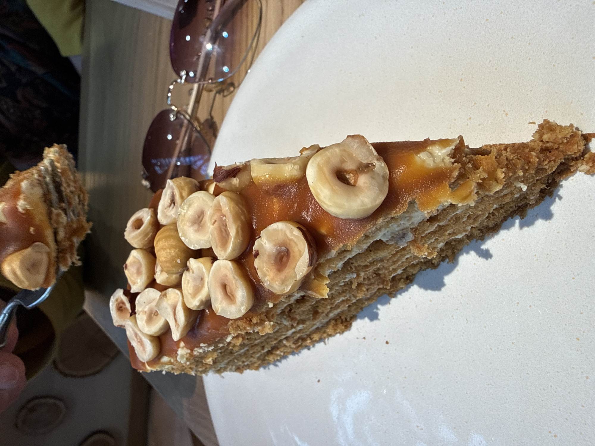 A close-up shot depicts a slice of layered cake topped with what appears to be nuts and a caramel-like glaze on a white plate. To the left of the cake are brown sunglasses. The location is Перепечино, Russia.