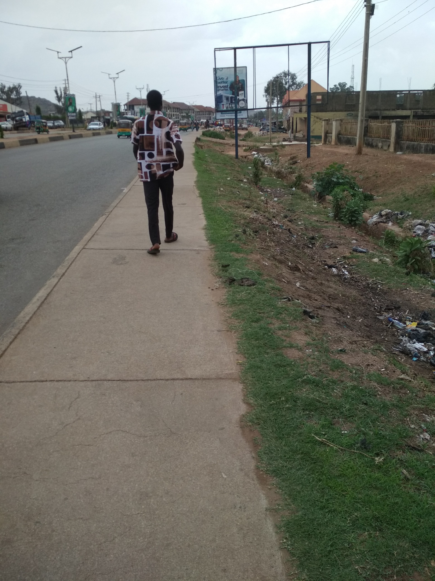 A medium shot captures a person walking away from the camera on a gray concrete sidewalk. The person is wearing brown trousers and a short-sleeved shirt with a collage pattern of brown, white, and black rectangles. Their dark hair is visible above the shirt. The sidewalk runs parallel to a road on the left, with vehicles like cars and tuk-tuks visible in the distance.
To the right of the sidewalk, there's a strip of green grass transitioning into a dirt patch littered with trash. In the background, a large rectangular billboard stands near a fenced area and some buildings. Power lines are visible against the overcast sky.