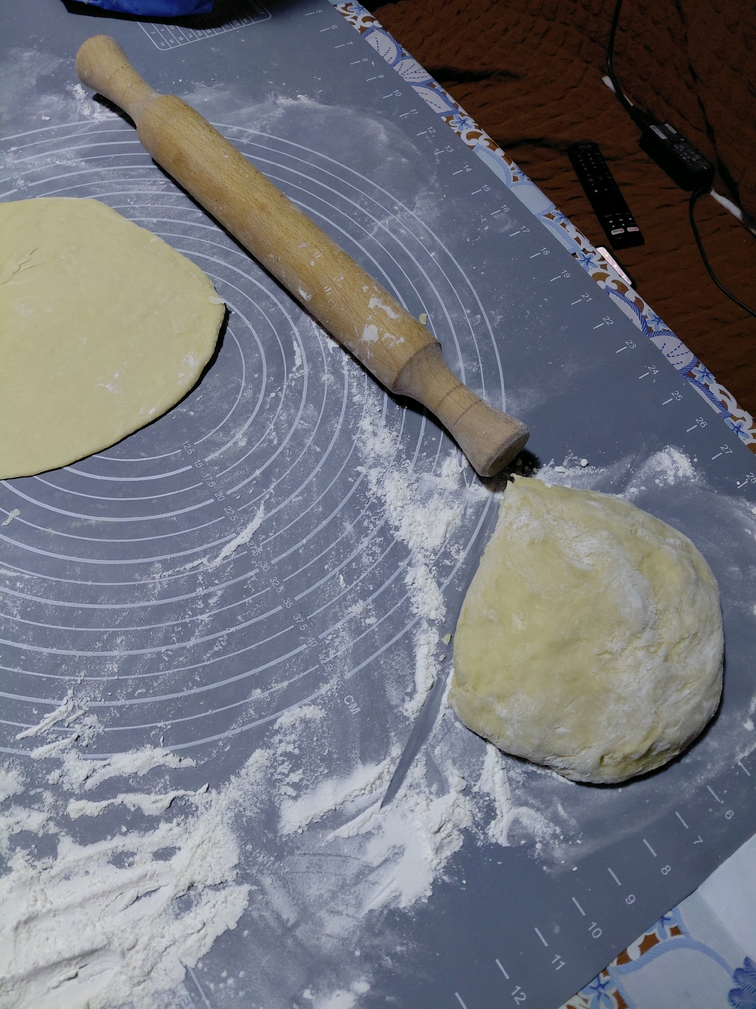 A close-up overhead shot shows a wooden rolling pin positioned on a gray silicone baking mat. To the left of the rolling pin is a portion of flattened dough, and to the right, a ball of dough is partially cut. Flour is dusted across the mat and the dough. The mat has circular guides and ruler markings along its edge. In the background, a patterned tablecloth and two remote controls are visible. The scene is located in an indoor environment.