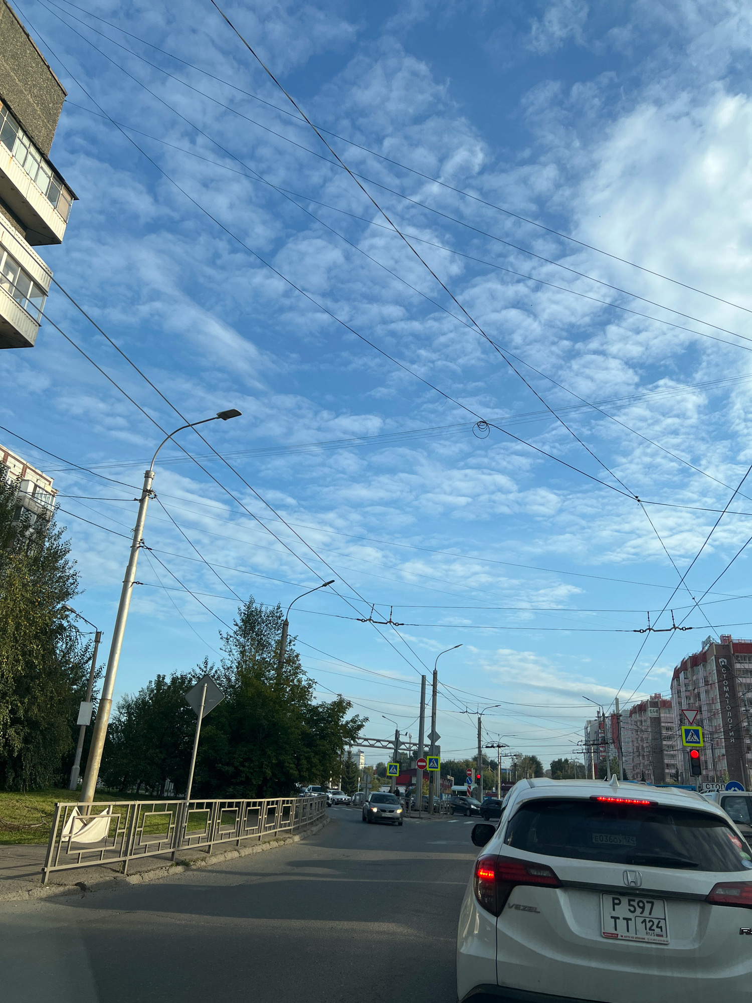 The image shows a street scene in Krasnoyarsk, Russia, on a partly cloudy day. The foreground features the rear of a white Honda Vezel with a Russian license plate "P 597 TT 124". The car is in traffic, as indicated by the red traffic light visible on the right side of the image. Alongside the road, there is a metal fence with square openings, and beyond that, trees and buildings. Overhead, numerous power lines and trolleybus wires crisscross the sky, supported by poles. In the background, more buildings and traffic are visible. The sky is blue with scattered white clouds.
