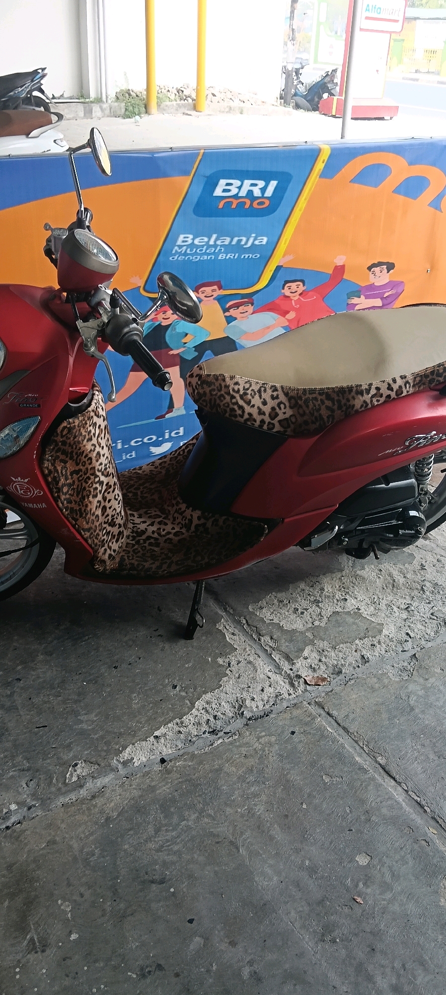 A red Yamaha Mio scooter is parked on a concrete surface in front of a banner that advertises BRImo, a mobile banking service. The banner has cartoon depictions of people and the text "Belanja Mudah dengan BRImo". Part of an Alfamart store sign is visible in the background. The scooter has a leopard print seat cover.