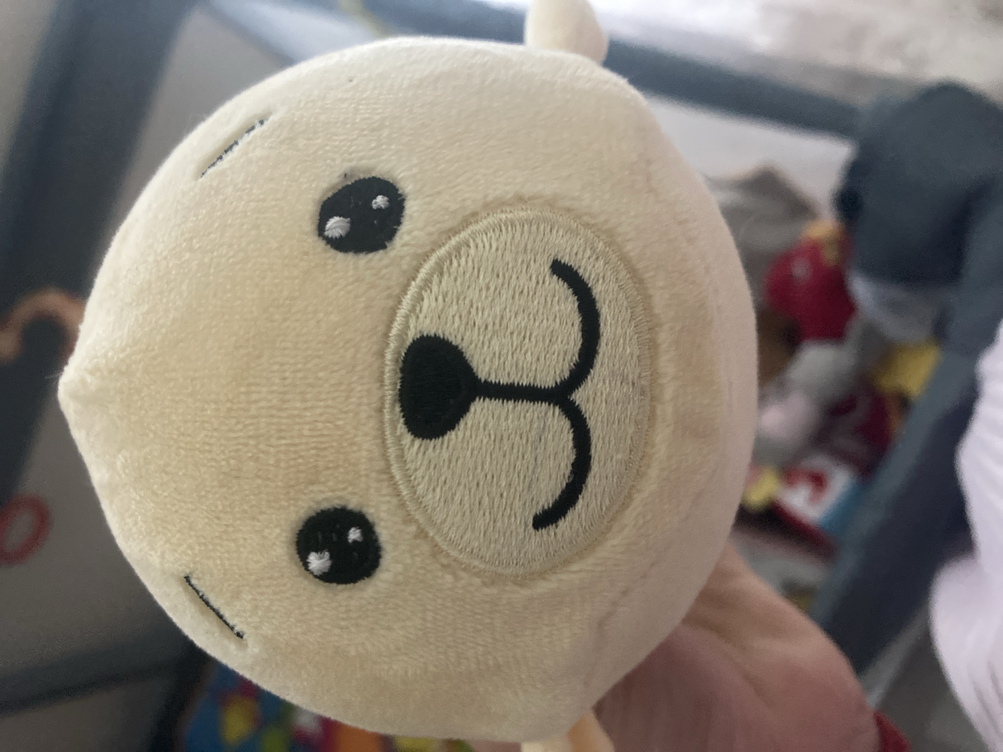 A close-up, head-on shot features a light tan, plush seal toy being held by a person whose hand is visible on the left side of the frame. The seal has black embroidered eyes with white highlights and a black embroidered smile. The background is out of focus and appears to be an indoor setting with colorful toys and other objects. The image was taken in Burgas, Bulgaria.
