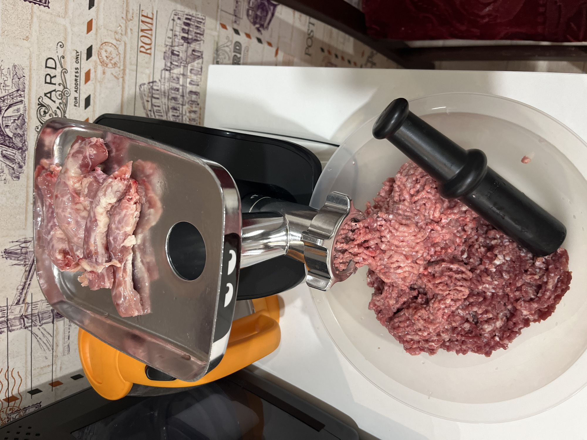 The image shows a meat grinder in operation. Raw meat pieces are in the silver feeding tray of the grinder. Ground meat is accumulating in a white plastic bowl next to the grinder's outlet. A black plastic food pusher is resting in the bowl with the ground meat. The grinder is placed on a white surface. The background consists of a patterned surface suggesting a countertop or backsplash with images related to travel, and part of a chair is also visible. There is no indication of the location being Chaikovsky, Russia.