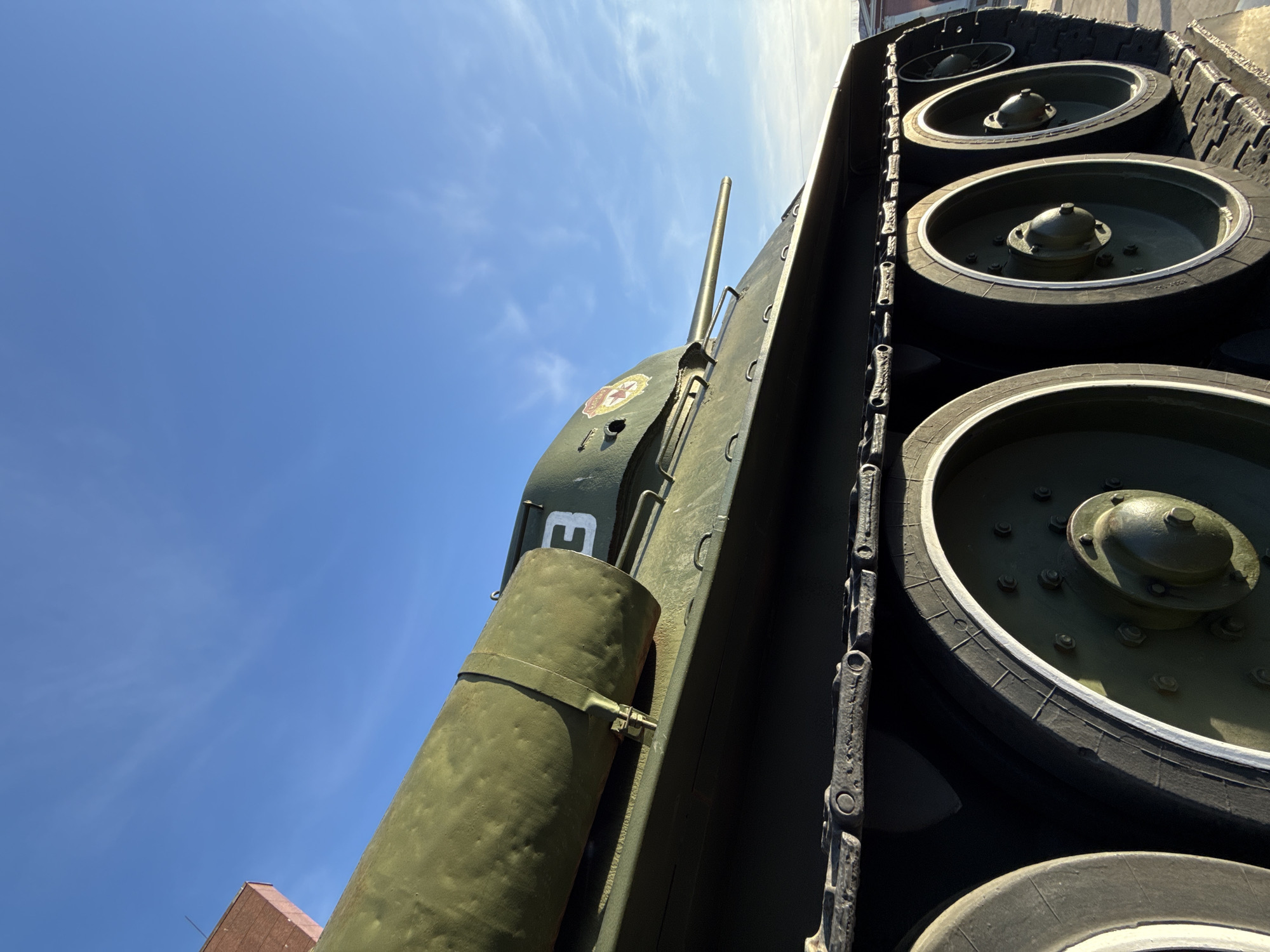 A forensically described image depicts an olive-green Soviet T-34 medium tank positioned outdoors under a clear blue sky with scattered cirrus clouds. The tank's right side dominates the frame, showcasing its track links and four visible road wheels with rubber tires and green metal hubs. A main gun barrel protrudes from the turret, which is adorned with a red star emblem featuring a golden outline and a hammer and sickle symbol. A partial white numeral "3" is visible on the turret. An auxiliary olive-green fuel drum is strapped horizontally to the tank's hull. Direct sunlight illuminates the scene, casting defined shadows. In the upper left background, a portion of a reddish-brown brick building is visible. There are no individuals, active movements, or interactions depicted. This scene is consistent with a military monument or museum exhibit, and in Perm, Russia, such a display would likely be found at a public memorial, a park, or a military history museum like the Perm Museum of Military History.