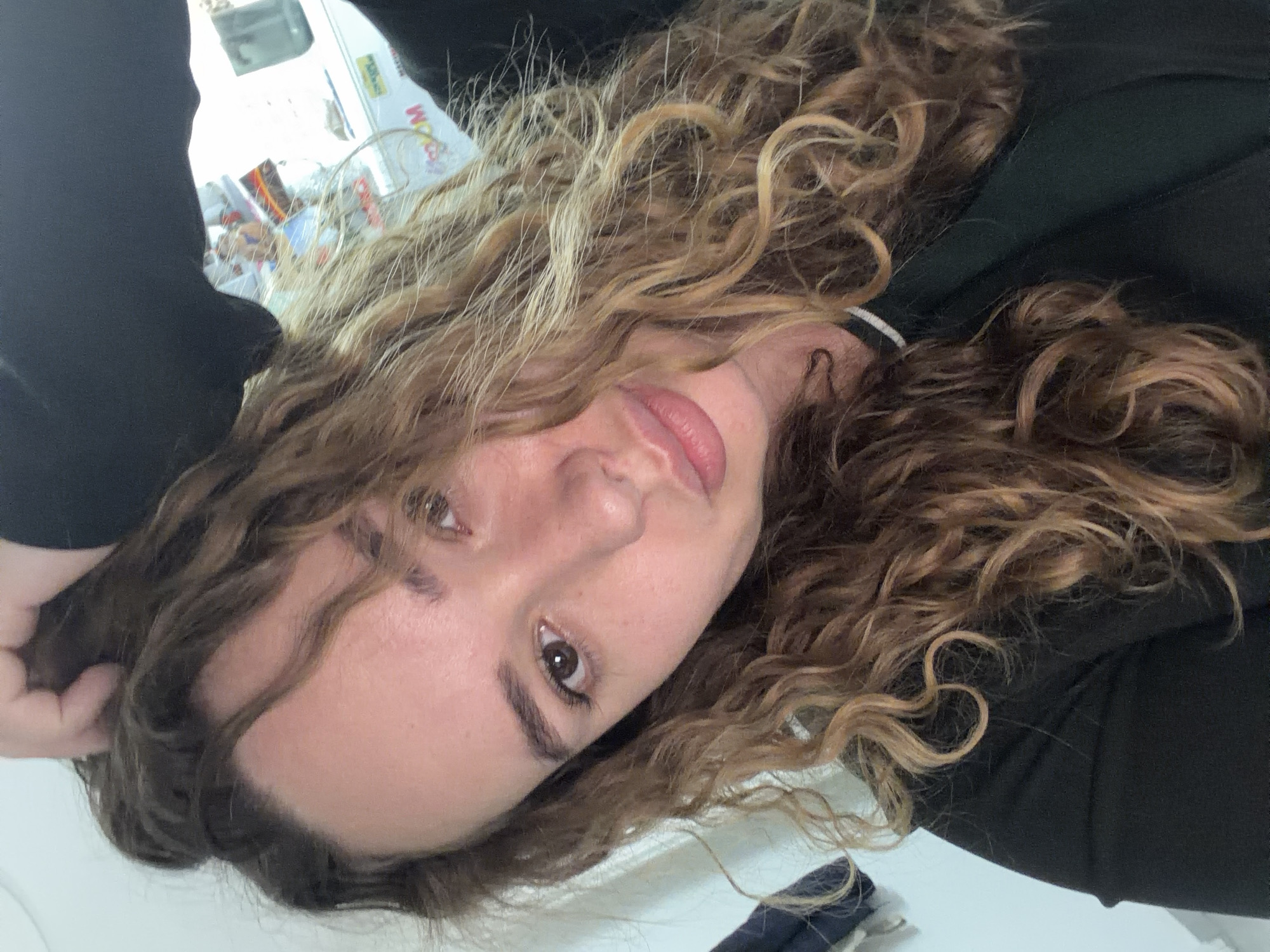 A woman with curly blonde and brown hair is looking at the camera. She has light skin, brown eyes, and full lips. She is wearing a black top. A white van with colorful stickers is visible in the blurred background, suggesting a location in Ramat Gan, Israel.