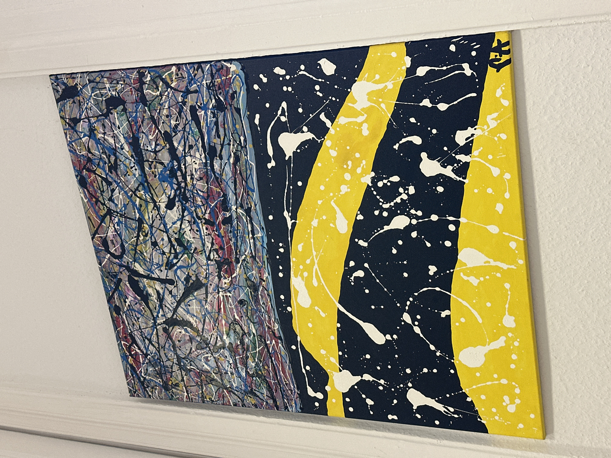 An abstract painting is prominently displayed on a white wall, with a white ceiling trim visible along the top edge of the frame. The artwork is composed of two distinct halves. The left side features a dense, multi-layered splatter and drip technique, with thin lines and dots of blue, white, black, red, and yellow paint creating a chaotic yet intricate pattern over a lighter background.
The right side of the painting contrasts sharply with a dark navy blue background. Across this dark base are three broad, slightly curved vertical stripes of bright yellow. White paint splatters and drips are scattered across this section, overlaying both the navy blue and yellow areas. In the top right corner of the painting, a small, stylized black symbol or signature is visible on one of the yellow stripes.
The painting is hung at a slight angle, appearing to tilt downwards to the right. The white wall has a visible, subtle texture. No people or specific activities are depicted in the image, which focuses solely on the artwork and its immediate surroundings.