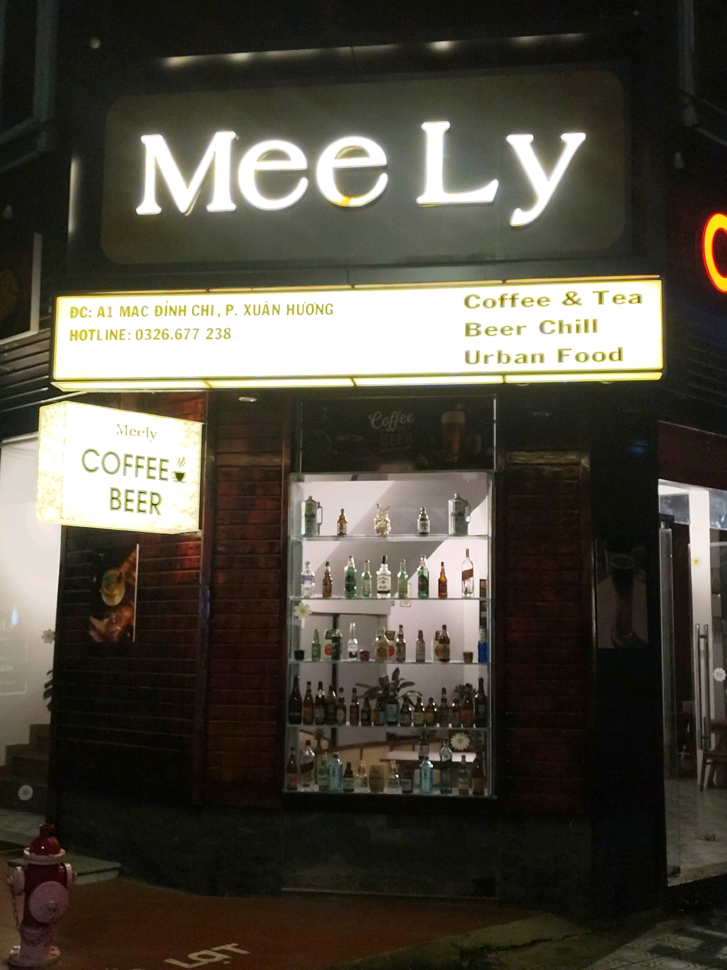 The image shows a nighttime street view of a business establishment in Da Lat, Vietnam. A prominent illuminated sign at the top reads "Mee Ly" in large white lettering. Below this, a rectangular illuminated sign contains text in yellow. The left side of this sign reads "DC: A1 MAC DINH CHI, P. XUAN HUONG" and "HOTLINE: 0326.677 238". The right side of this sign lists offerings: "Coffee & Tea", "Beer Chill", and "Urban Food".
To the left of the main entrance, a square, illuminated sign is suspended, displaying "Meely" at the top, followed by "COFFEE" and "BEER" with a small coffee cup icon next to "COFFEE". The entrance area features a brick facade.
In the center of the image, a glass-fronted display case showcases a collection of bottles and decorative items on multiple shelves. The bottles appear to be liquor or beer bottles of various sizes and colors.
On the lower left side of the image, a red and silver fire hydrant is visible on the sidewalk. The ground in front of the establishment is paved, with some visible cracks. The overall environment suggests an urban setting at night.
