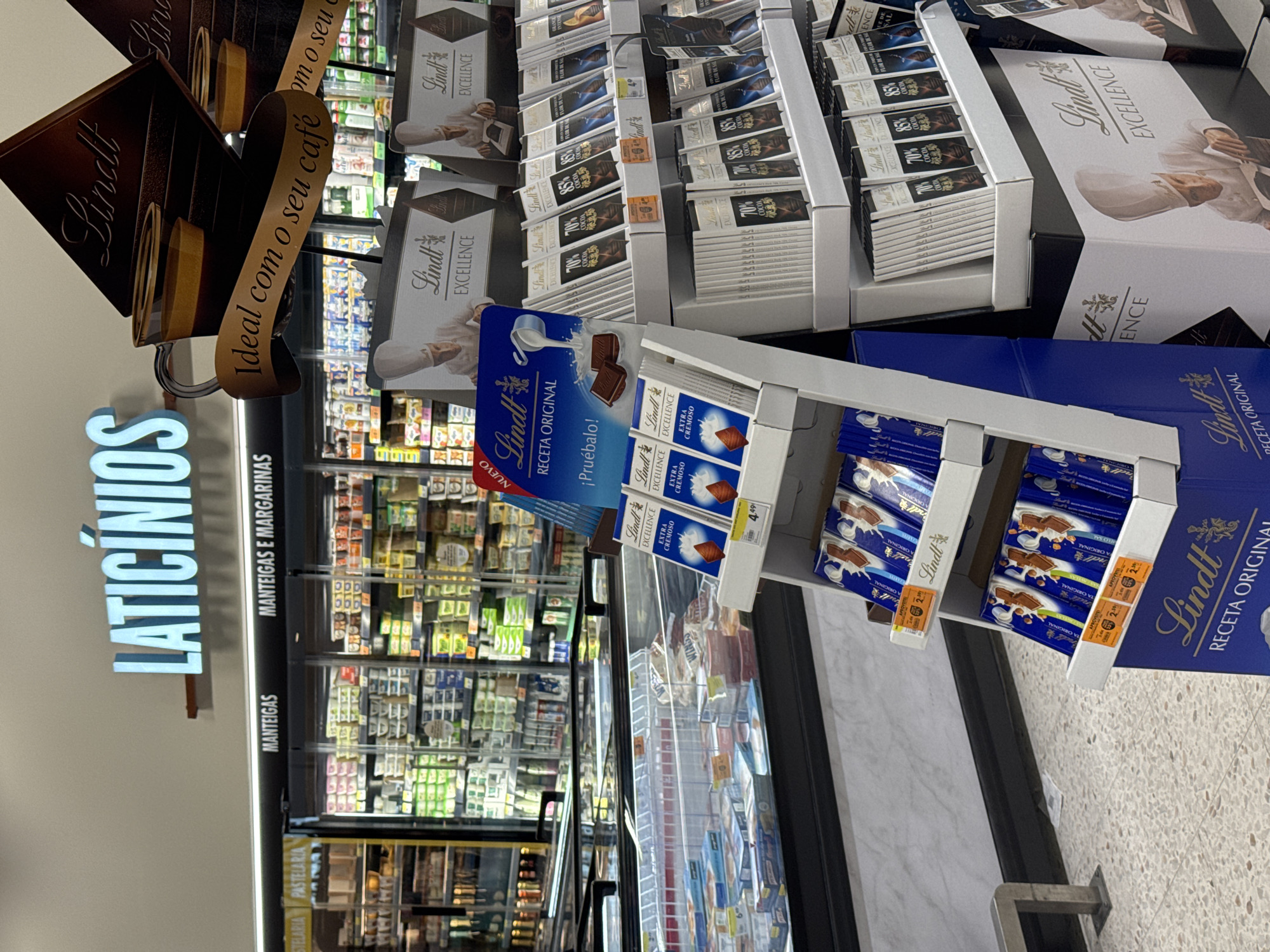 The image shows the interior of a supermarket, likely a Pingo Doce, as indicated by the signage and product placement. The overall scene is focused on a display of Lindt chocolate bars and other confectionery. In the background, refrigerators are filled with dairy products, and overhead signage reads "LATICÍNIOS" (Dairy Products) in large blue letters, and "MANTEIGAS" (Butters) and "MANTEIGAS E MARGARINAS" (Butters and Margarines) in smaller black letters. The promotional display features triangular cardboard cutouts of a coffee cup with the text "Ideal com o seu café" (Ideal with your coffee) in Portuguese. Below this, various Lindt chocolate bars, including "Excellence" and "Receta Original" lines, are neatly arranged on shelves. Some of the chocolate bars are priced, with tags showing prices like "4.49€" and "2.99€". In the foreground, a blue cardboard box displays more Lindt chocolate bars, emphasizing "Receta Original". The floor is tiled with a speckled pattern. The presence of Portuguese text on the signage and product packaging strongly suggests the location is within Portugal. Based on the specific branding and typical store layout, this supermarket is most likely located in Castelo Branco, Portugal. There are no people visible in the frame.