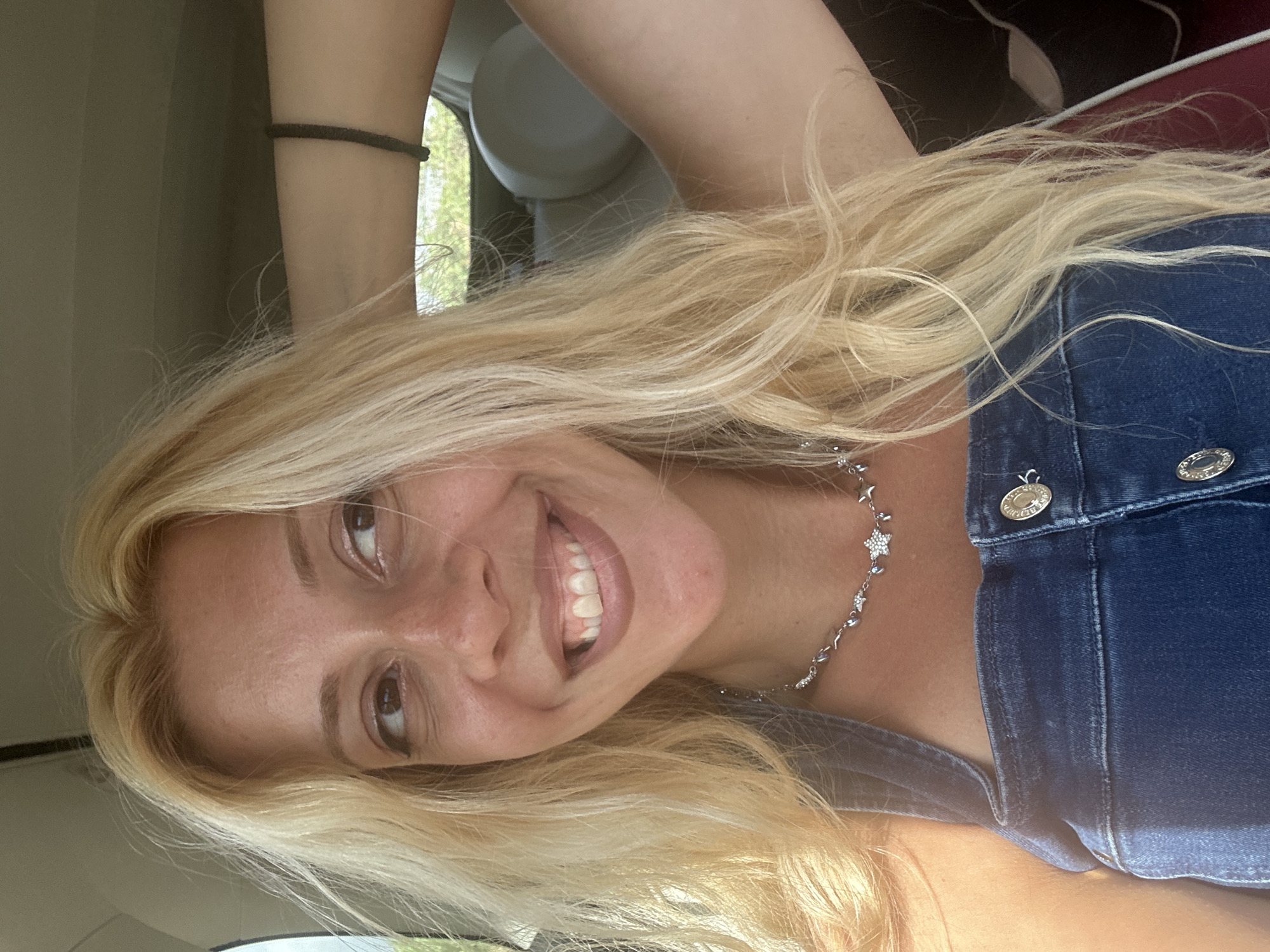 A young Caucasian woman with long, blonde, wavy hair and fair skin is captured in a selfie taken from inside a vehicle. She is wearing a dark denim overall with visible buttons and a delicate silver necklace with star-shaped charms. Her left arm is raised and resting on her head, while her right arm is also raised and bent, with her hand near her head. She has brown eyes and a wide, genuine smile, with her head tilted slightly to her left. The background suggests an outdoor environment with glimpses of greenery and a car's interior, including a headrest. The lighting indicates it is daytime, possibly sunny, with light casting shadows on her face and body. The image does not provide any information about the location being in Offin, France.