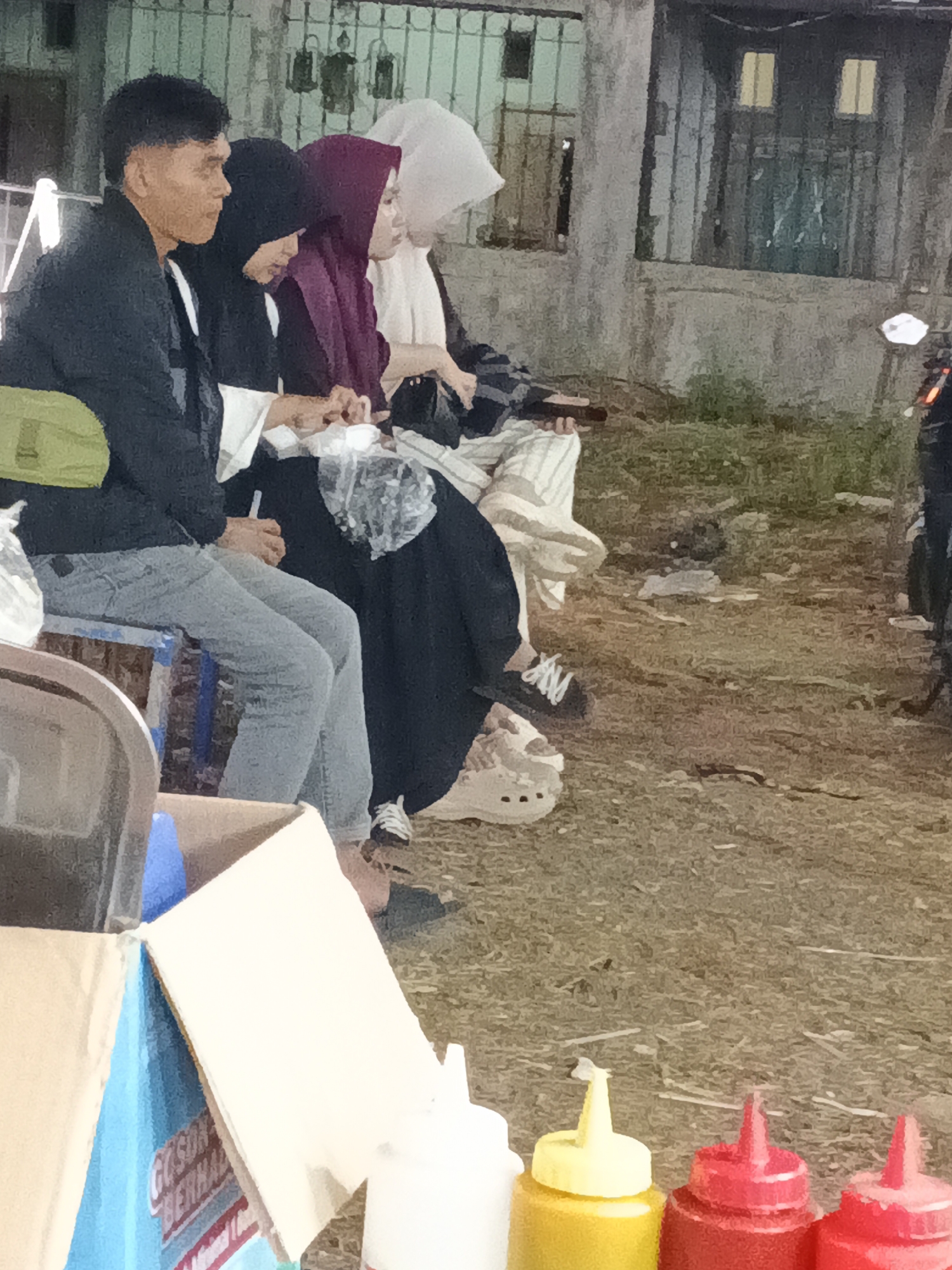 In Sukabumi, Indonesia, four individuals are seated outdoors on a gritty, uneven surface. A man in a dark jacket and light-colored pants is on the far left, with a green bag slung over his shoulder. Next to him, a woman wears a black hijab, followed by another woman in a maroon hijab. The person on the far right has a white head covering and is partially obscured. They appear to be engaged in a social gathering. In the foreground, a cardboard box contains what appear to be condiments, with at least three squeeze bottles visible: one yellow, one red, and another red one. A metal fence with vertical bars is in the background, and a building with barred windows can be seen behind it. The lighting suggests it is either dusk or nighttime.