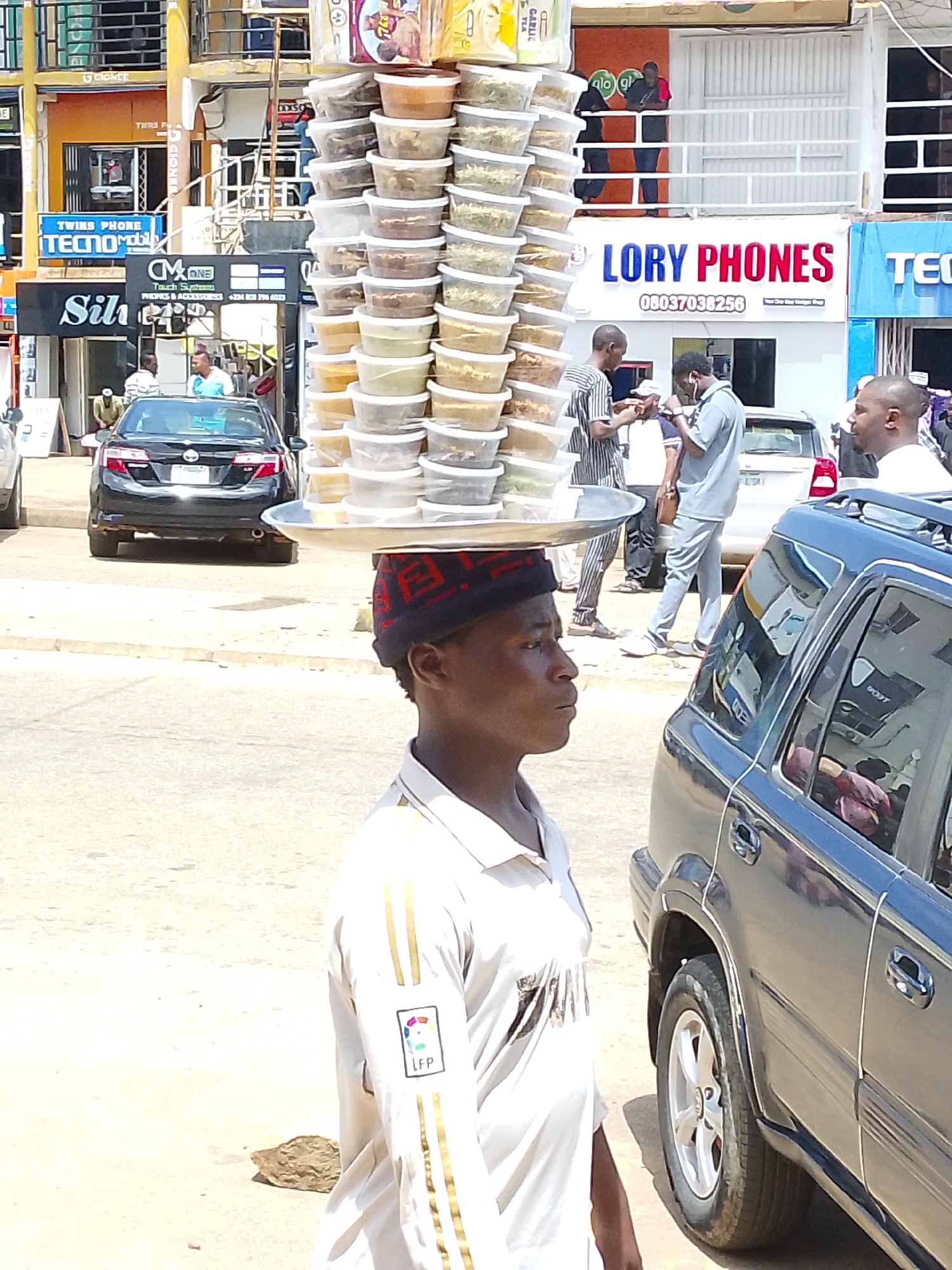 A man carries a tall stack of plastic food containers balanced on his head in a street scene in Jos, Nigeria. The containers, filled with various food items, are arranged in columns on a metal tray. The man, wearing a light-colored polo shirt with gold stripes and a patterned cap, is in profile, facing right. In the background, several vehicles are visible, including a black sedan and a dark SUV. Several people are also present, some standing and interacting, while others appear to be customers or passersby. Buildings with signage for businesses like "LORRY PHONES" and "TWINS PHONE" are seen lining the street, suggesting a commercial area. The environment appears to be an outdoor, sunny urban setting.