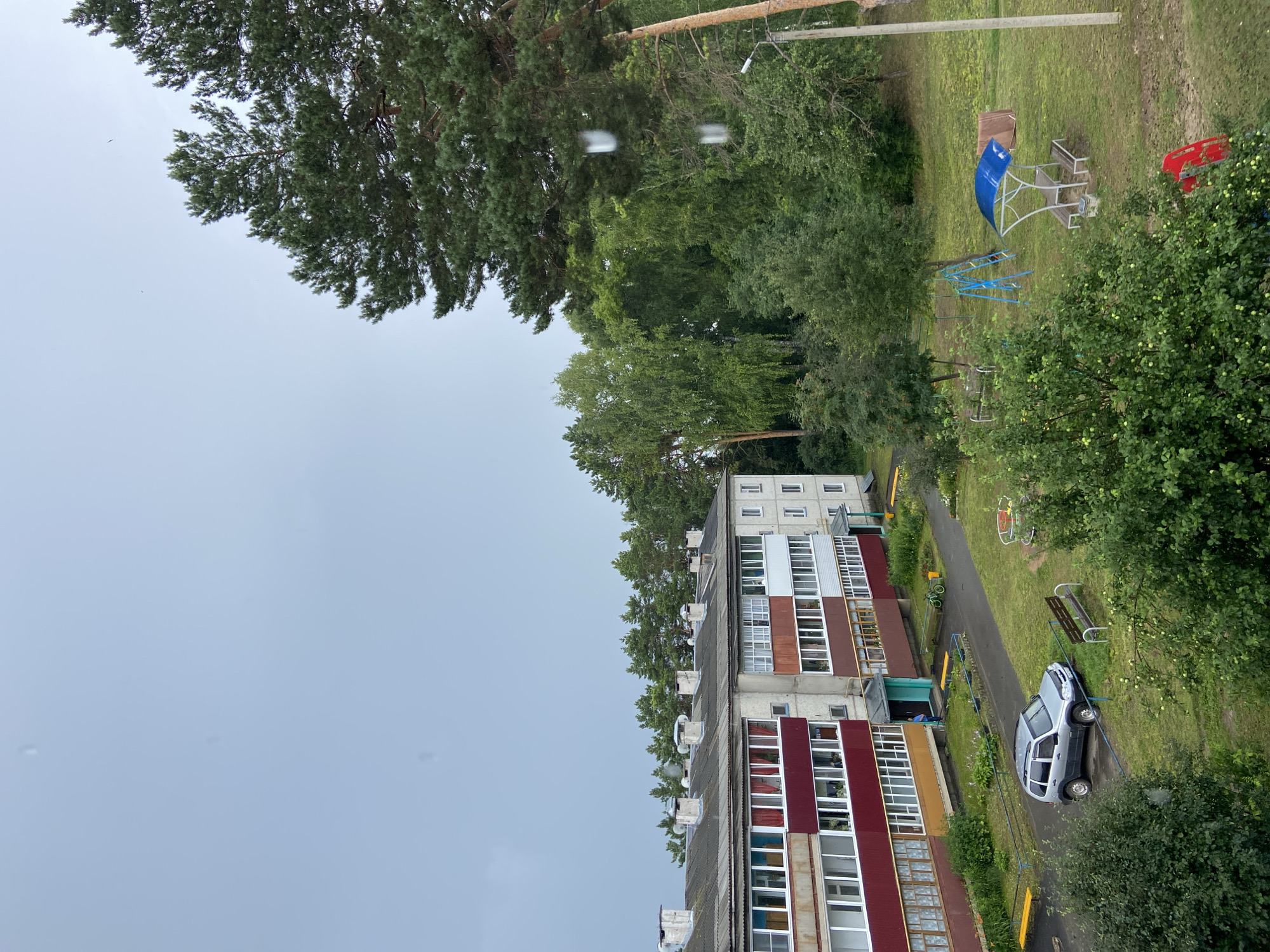 The image depicts a courtyard area in a residential complex. In the foreground, a silver SUV is parked on a grassy area next to a dark asphalt path. Further back, a light gray, multi-story apartment building with red and brown balconies is visible. A playground area with a blue slide and a canopy with a picnic table is located in the middle ground. Dense green trees and vegetation surround the scene, and the sky is overcast and light gray. There is a single person standing near the entrance of the apartment building. The overall impression is that of a utilitarian, common residential area in a possibly rural or suburban setting, likely experiencing cloudy or rainy weather.