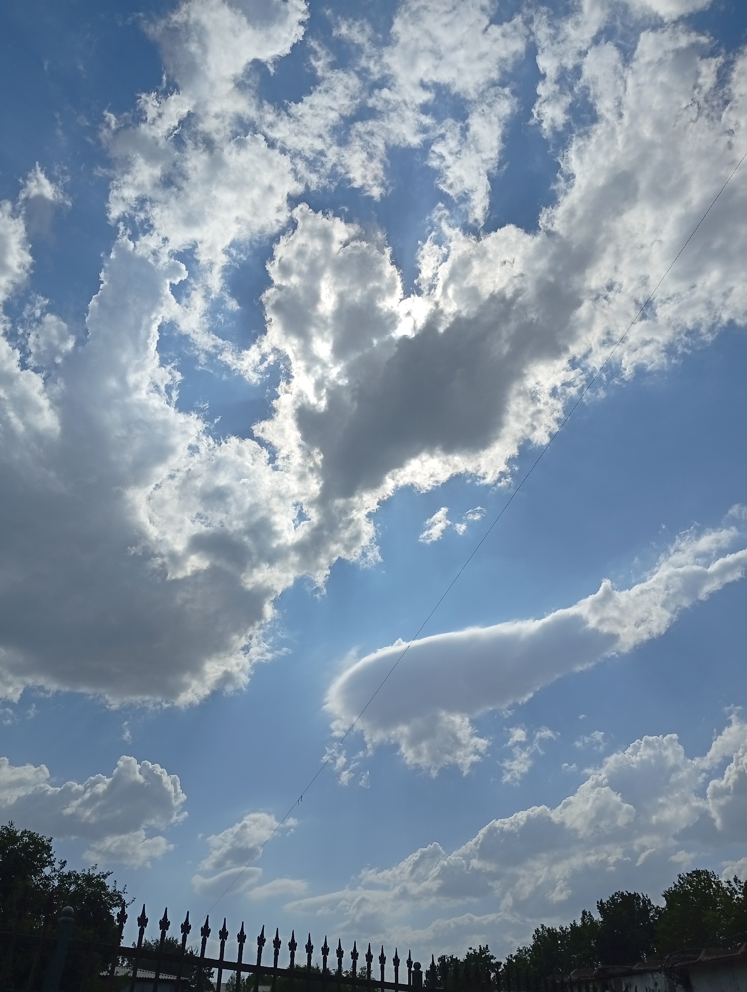 The image captures a sky filled with dynamic clouds, showcasing both bright white formations and darker, shadowed areas, suggesting a mix of sunlight and impending weather. The blue sky peeks through in various patches, creating a dramatic contrast. A thin, dark wire stretches diagonally across the frame from the upper right to the lower left, possibly a power line or telephone wire. In the lower portion of the image, a dark metal fence with pointed finials runs horizontally. Behind the fence, the tops of trees are visible, with dark green foliage suggesting an outdoor environment. The overall scene depicts a sky view over a partially obscured landscape, characteristic of a city or suburban area. The location is specified as Angren, Uzbekistan.