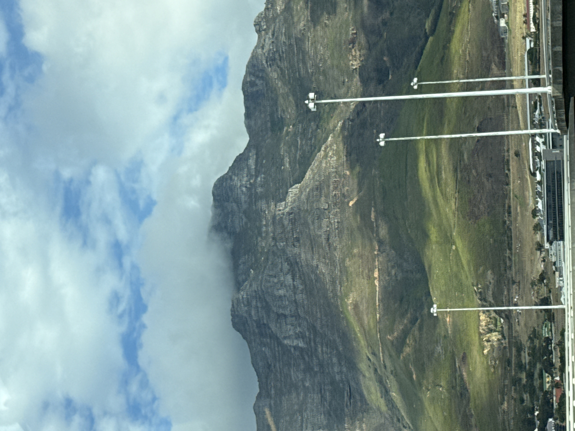 The image shows Table Mountain in Cape Town, South Africa. The mountain's rocky face is visible, partially obscured by clouds. Green vegetation covers some areas. Several tall light posts line the right side of the frame, extending into a cityscape in the distance. The sky is blue with white clouds.