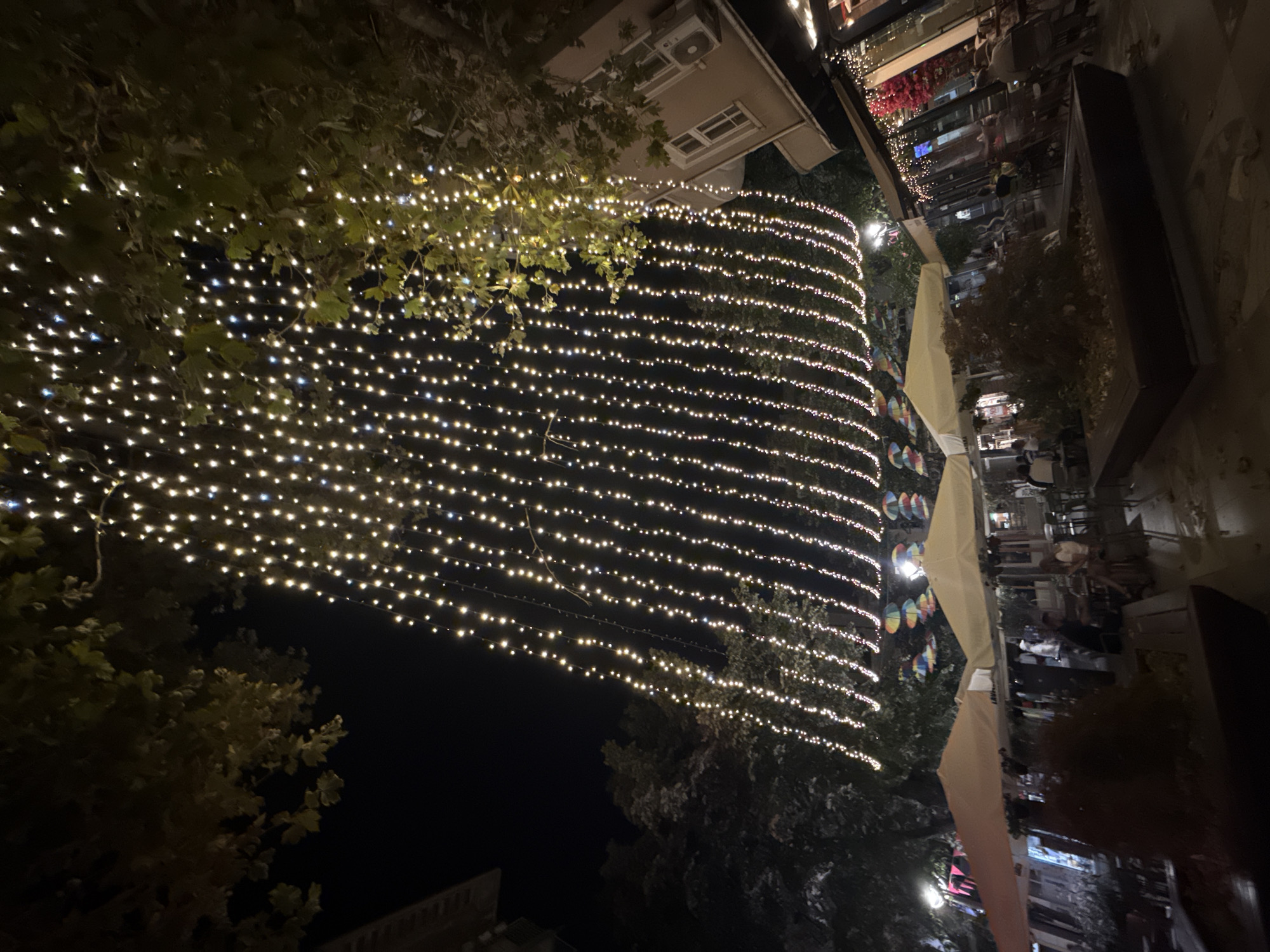 The image depicts a nighttime scene in the city of Burgas, Bulgaria, likely on a pedestrian street or promenade, illuminated by numerous strands of fairy lights strung overhead. The lights create a cascading effect against the dark night sky, with trees whose leaves are interspersed among the glowing strands. Below the lights, several outdoor seating areas for cafes or restaurants are visible. Two large, cream-colored umbrellas are open, providing shade. In the background, buildings with windows and air conditioning units can be seen. Some people are seated at tables, engaged in conversation or dining. A few colorful umbrellas are also visible hanging in the distance. The overall ambiance is festive and inviting, suggesting a lively urban setting.