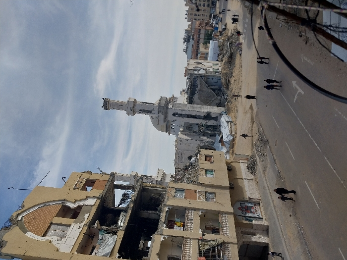 This image depicts a scene of widespread urban destruction in Khan Yunis, Palestinian Territory, under a partly cloudy sky.
In the left foreground, a multi-story building, likely residential or commercial, is severely damaged. Its light-colored facade is partially torn away, exposing internal structures, rebar, and debris within multiple floors. Balconies are compromised, and window openings are visible but largely empty.
Centered in the mid-ground, a large mosque-like structure exhibits extensive damage, with a distinctive dome and a tall, partially collapsed minaret that leans slightly. Large sections of the mosque's walls are missing, revealing interior spaces, and significant amounts of rubble surround its base. Adjacent to the mosque, other structures are reduced to rubble and debris.
The paved street in the foreground features white lane markings, including a solid line and a turning arrow, and appears dusty or sandy. Several individuals are present on the street and amidst the debris. On the left side of the street, a man walks with a child. Multiple other pedestrians, dressed in civilian clothing, are scattered along the street and near the rubble piles further into the scene, with some forming small groups. The overall environment conveys a scene of significant devastation and ongoing activity by a few residents.