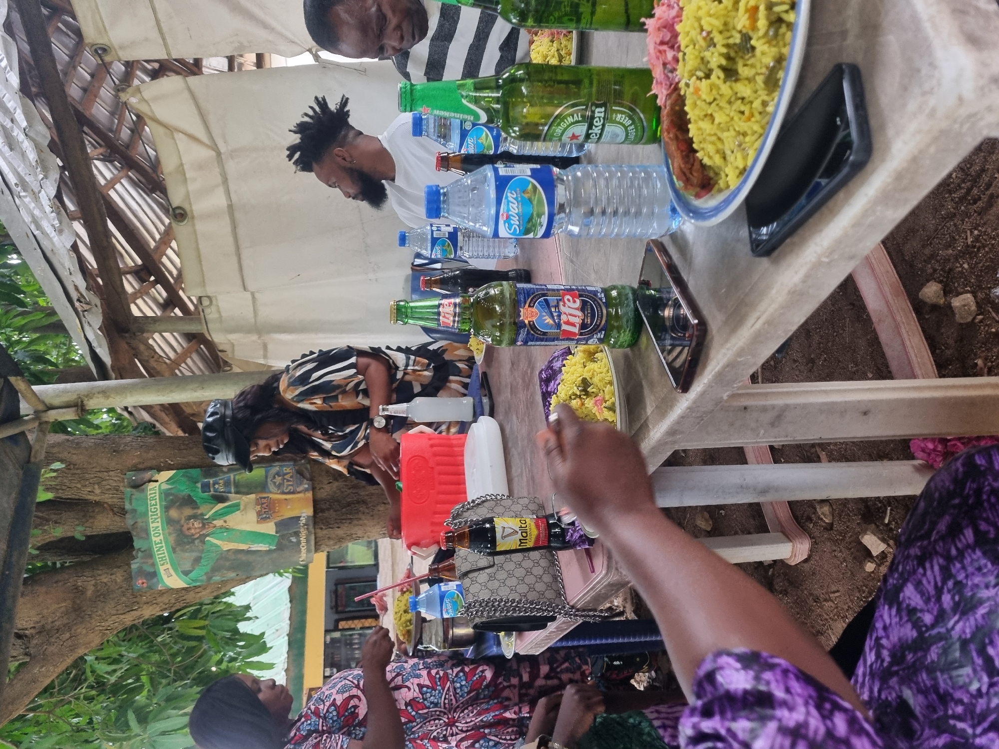 The image depicts a social gathering in what appears to be an outdoor setting in Jos, Nigeria. Several people are seated around a white plastic table laden with food and beverages. Food items visible include a plate of yellow rice with different colored toppings, and at least one other plate with food in a bowl. Beverages consist of bottles of Heineken and Life lager, Swan bottled water, and Malta Guinness.
The individuals present consist of men and women of varying skin tones and attires. The table appears to be located under a temporary shelter constructed from corrugated metal and white fabric. A sign for Star beer featuring the slogan "Shine on Nigeria" is attached to a tree nearby. Other visible objects include a red cooler, a handbag with a chain strap, and a smartphone. The ground beneath the table is dirt.