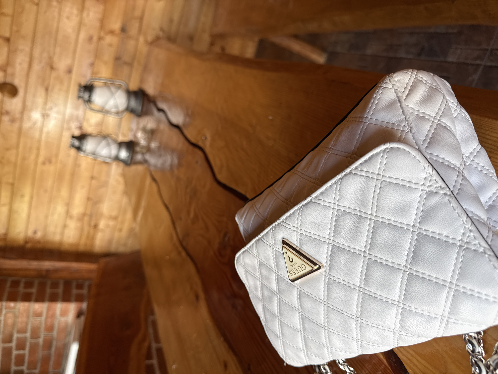 A white quilted handbag with a gold "Guess" logo is displayed on a polished wooden table in a rustic interior setting. The table has a natural, unfinished edge and is situated in what appears to be a log cabin or a similar wooden structure, evidenced by the wooden paneling on the wall behind it. In the background, two vintage-style lanterns are placed on the table, one slightly behind the other. To the left, a section of a brick wall is visible, and a wooden bench is partially in view. The lighting suggests an indoor environment, possibly during the day, with a warm, ambient glow. The overall scene evokes a cozy, slightly antiquated atmosphere. The location is identified as Cârțișoara, Romania.