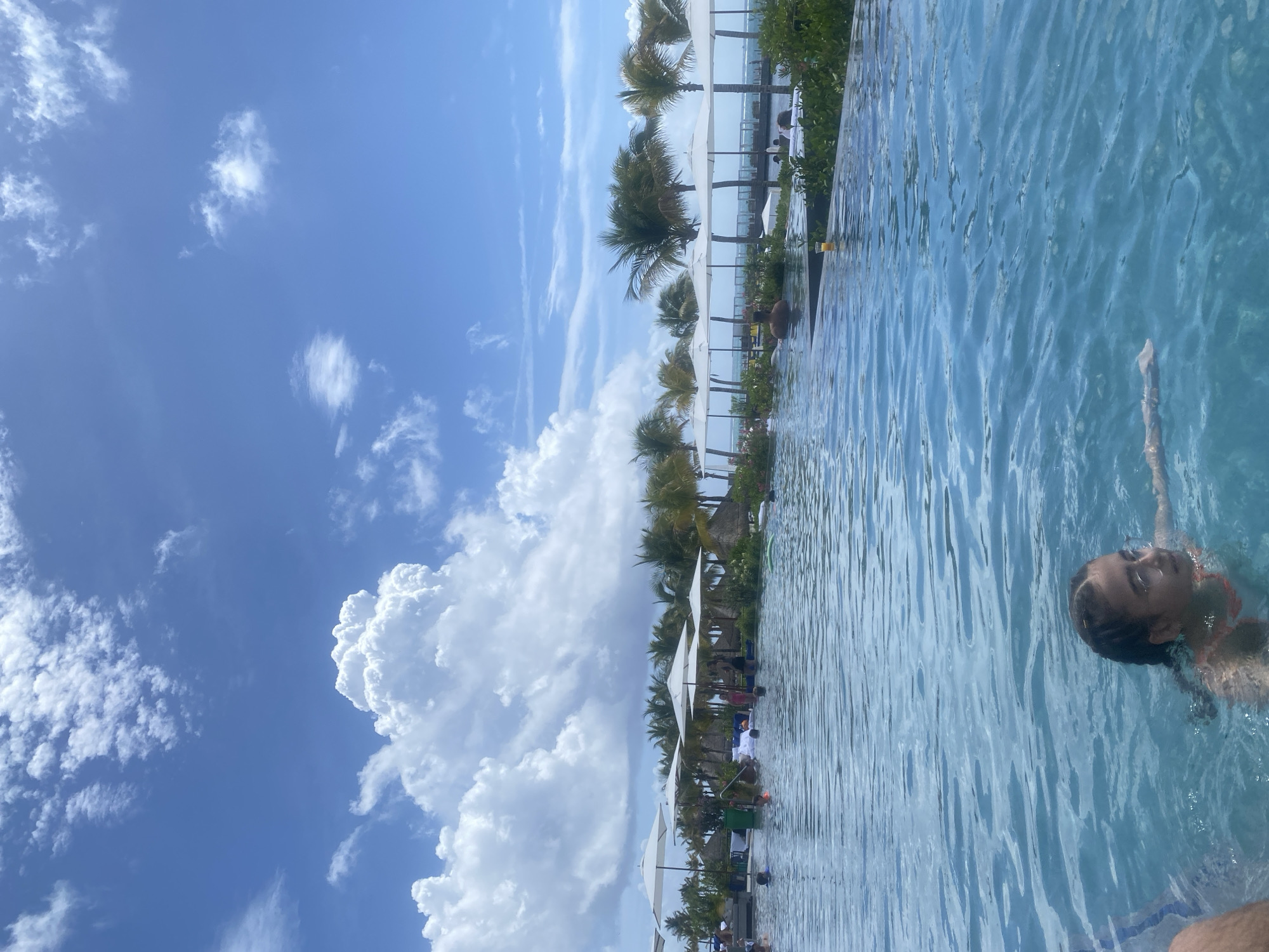 Here's a detailed description of the image:
**Location:** Marina Maroma, Mexico. The image appears to be taken at a resort featuring an infinity pool.
**Environment:** The sky is bright blue with large cumulus clouds. There are palm trees and white sun umbrellas lining the edge of the pool. The pool water is clear and light blue.
**People:** A person is swimming in the pool in the bottom right corner of the image. The person appears to be female. The pool edge is lined with sun umbrellas and beach chairs where several people are seated.
**Objects:** The main objects are an infinity pool, umbrellas, palm trees, chairs, and clouds.