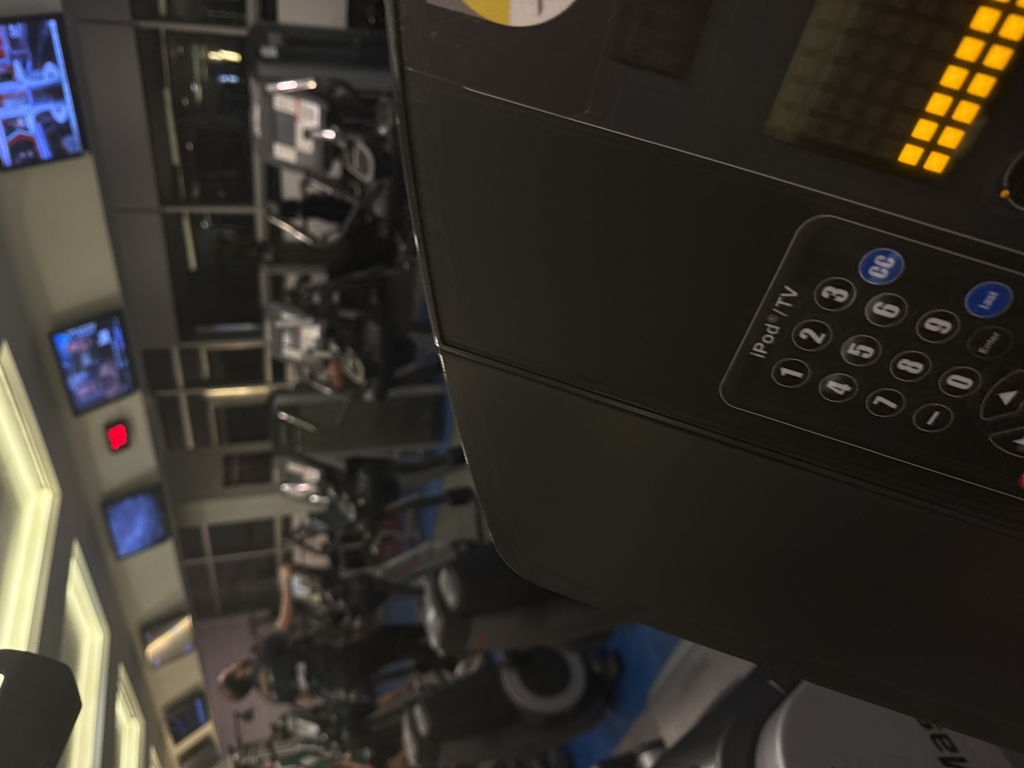 The image appears to be taken inside a gym. In the foreground, there's a close-up view of a treadmill's console with a keypad featuring buttons for numbers 0-9, CC (closed captions), and functions related to iPod/TV control. In the background, further into the gym, rows of exercise machines can be seen, some of which appear to be treadmills and stationary bikes, occupied by individuals working out. On the wall, there are several televisions displaying various content. A red digital clock is also visible on the wall. The gym appears to be indoors, possibly in Mexico City based on the prompt's instruction. The environment is brightly lit, suggesting standard gym lighting.