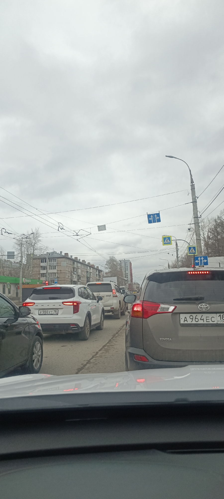 The image depicts a traffic jam in the city of Izhevsk, Russia. Several vehicles are stopped on the road, including a Toyota RAV4 with license plate A964EC 18 RUS, a white Haval F7x with license plate P988YT 18, and other SUVs. Buildings can be seen in the background, and street signs indicating pedestrian crossings and traffic directions are present. Overhead power lines and a cloudy sky are also visible. The perspective is from inside a car, looking out through the windshield.