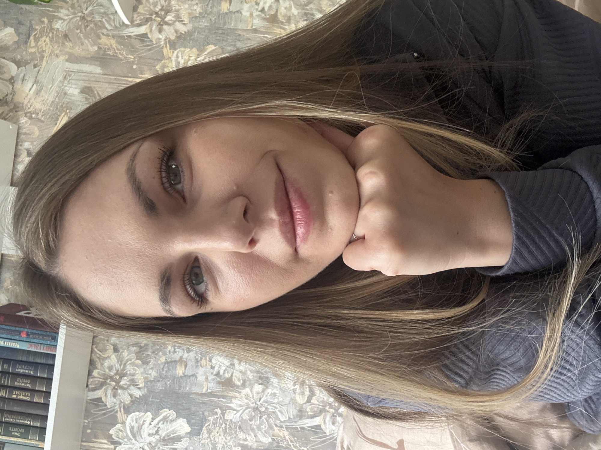 A woman is pictured indoors with her head resting on her hand. She has long blonde hair and fair skin. She is wearing a dark blue or gray ribbed top. The background includes a bookshelf filled with books and wallpaper with a floral design. The location is Раздолье, Russia.