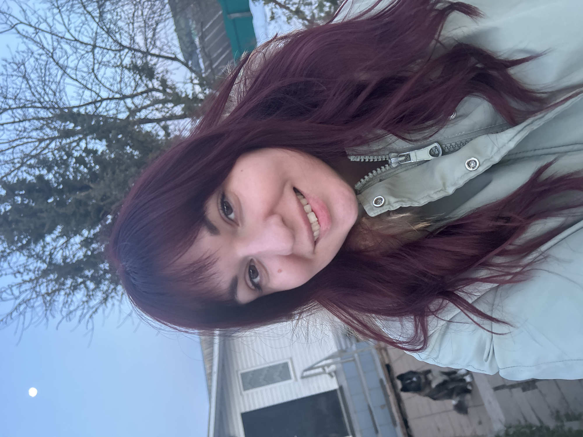 A woman with burgundy hair and wearing a light green jacket is smiling in a selfie-style photo. Bare tree branches are visible behind her in the upper left corner, while the sky is a light blue, possibly suggesting dusk or dawn. A white house is visible in the lower background. A dog can be spotted sitting behind the house, to the right. The location is in Фоки, Russia.