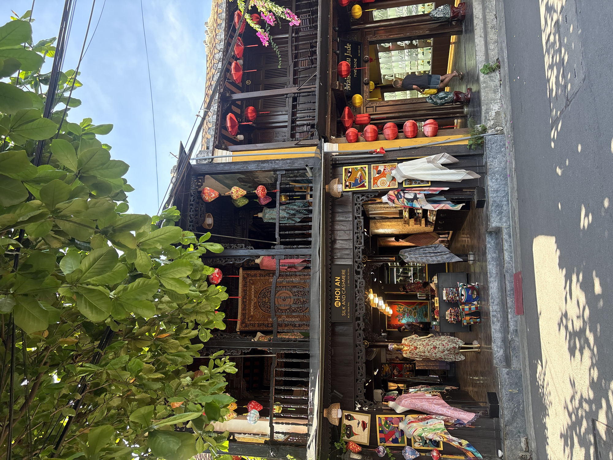 The image captures a street view of a shop in Hội An Ward, Vietnam. The shop, named "HOI AN SILK AND CASHMERE", is two stories high with dark wooden balconies. It displays a variety of goods, including mannequins dressed in colorful scarves and clothing, artwork, and textiles hanging on the walls. Red lanterns are strung along the balconies and building facade, adding a festive touch. To the right, another shop, "Maison du Jade", is visible, also adorned with red lanterns. A person wearing a dark t-shirt and shorts is seen walking into Maison du Jade. The street in front of the shops is paved, with dappled sunlight casting shadows from overhead trees. The sky is clear blue.