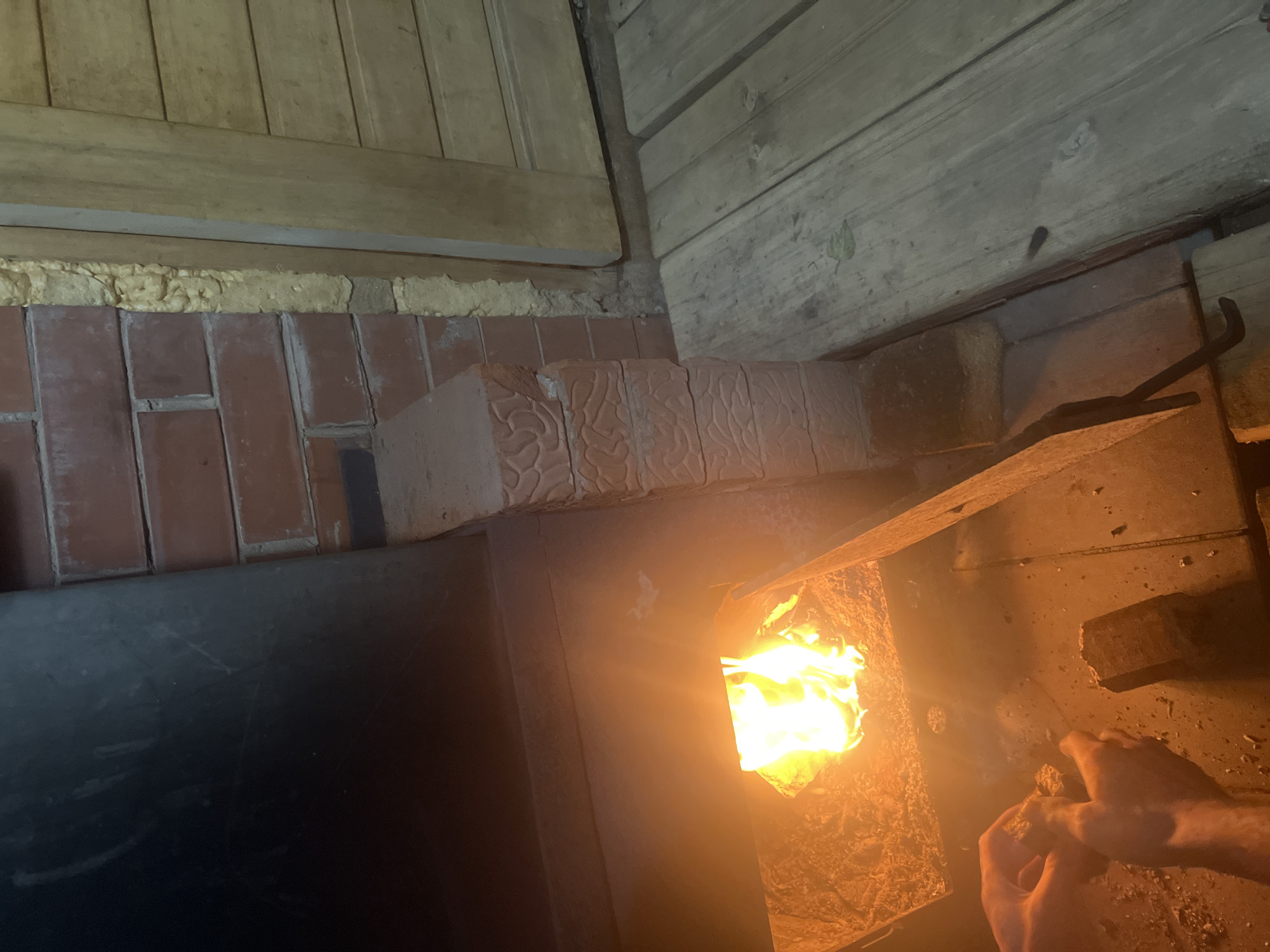 The image shows a fireplace with flames inside. The fireplace is constructed of bricks and is located in a room with wooden walls and ceiling. A pair of hands are visible near the opening of the fireplace, holding something unidentifiable. The location is unknown and not indicated in the image. There is no evidence this image was taken in the city of Chaikovsky, Russia.