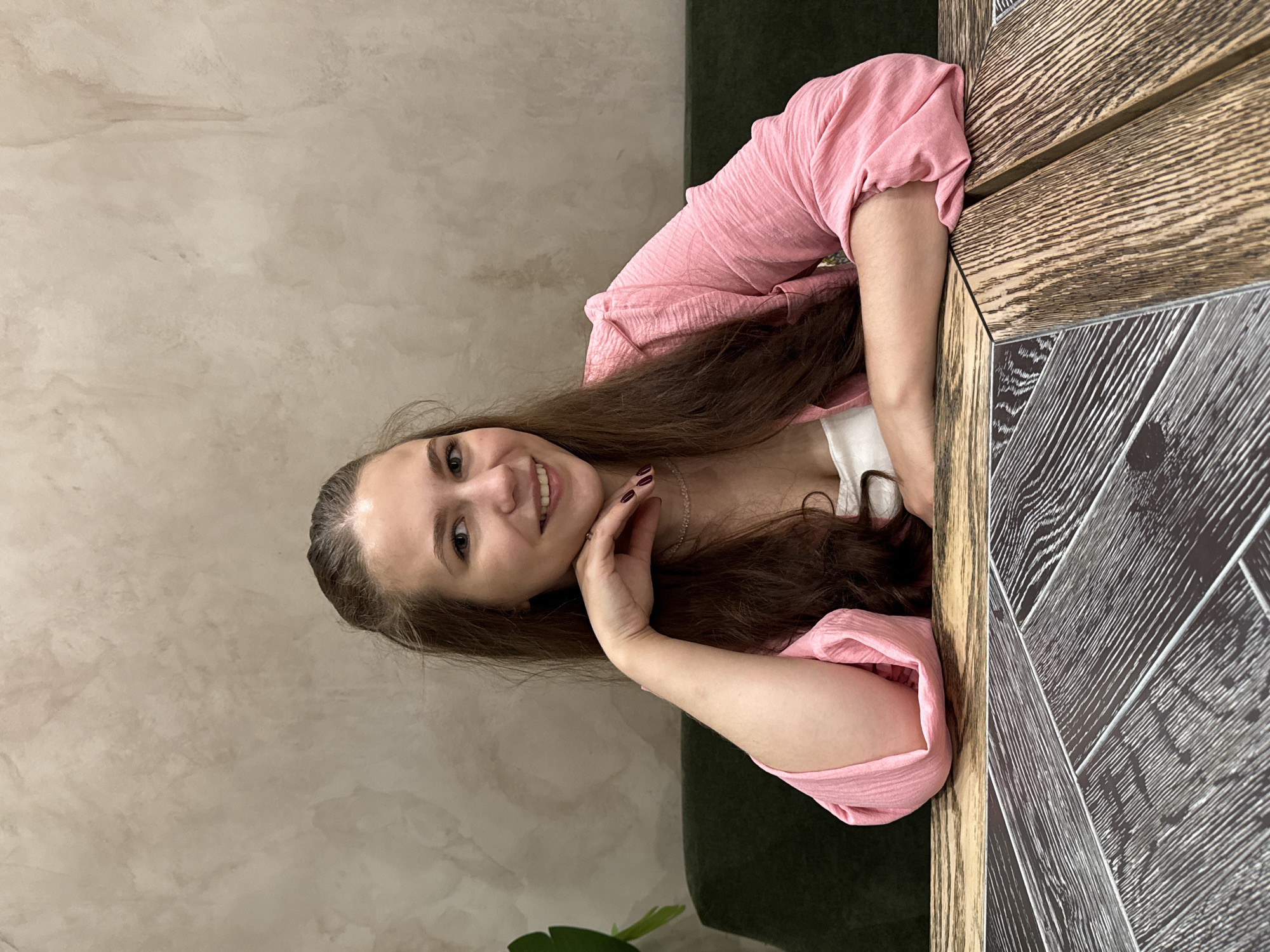The image depicts a young Caucasian woman with long, brown hair, seated at a table in what appears to be a restaurant or cafe. She is wearing a pink, textured jacket over a white top and a delicate necklace. Her right arm is resting on the table, with her hand near her chin, and she is smiling directly at the camera. The background shows a textured wall in a neutral tone and a dark green banquette seating. The table in the foreground has a distinctive pattern of dark and light wood. The image does not contain any specific visual cues to indicate its location within the city of Izhevsk, Russia.