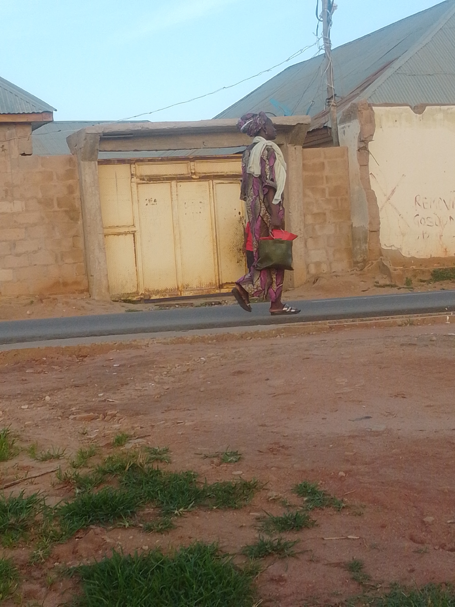 A full-body, side profile shot captures a person walking on a paved road in Gombe, Nigeria. The individual, dressed in a purple and pink patterned traditional outfit with a light-colored headscarf and a shawl draped over their shoulders, is carrying a green bag over their left shoulder. They are wearing sandals. Behind the person, a weathered yellow gate is set into a brick wall. The ground in the foreground is a mixture of reddish-brown earth and sparse green vegetation. In the background, a corrugated metal roof of a building is visible against a pale sky. The scene appears to be in an urban or semi-urban environment within Gombe.