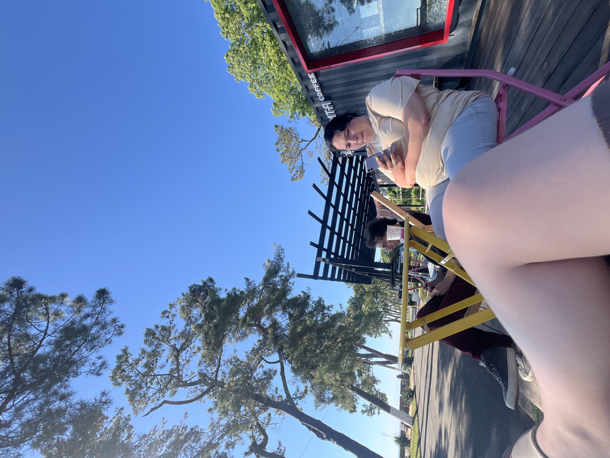 A person is sitting at a table outside a coffee shop. The person is holding a phone in their hand and looking at it. The coffee shop appears to be made out of a shipping container, with a red trim around a window. There is a tree with green leaves behind the coffee shop. The sky is blue, indicating a clear day. The sun is shining, casting shadows on the wooden deck where the table and chairs are located.
The person is wearing a light-colored t-shirt and light-colored pants. They have dark hair. The table is yellow, and the chairs are pink and yellow. A black fence is behind the table.
The image appears to be taken in the daytime, likely late morning or early afternoon, given the bright sunlight and clear sky. The setting appears to be an outdoor seating area for a coffee shop. There is visible text on the side of the shipping container that reads "COFFEE". The overall mood is relaxed and casual, with the person engrossed in their phone.