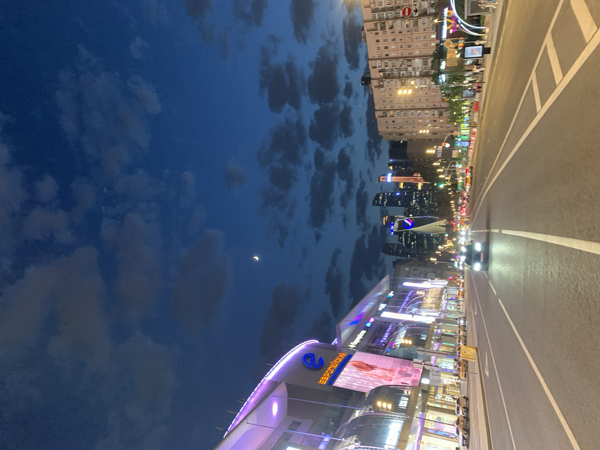 Captured at an unknown location in an unknown city, this image portrays a nighttime urban scene. The sky, occupying the left half of the frame, displays a dark blue expanse with interspersed clouds and a crescent moon. On the right, a multi-lane road stretches into the distance, illuminated by streetlights and headlights of vehicles. Buildings are arrayed on either side of the road, displaying a mix of architectural styles and illuminated facades. One building, distinguished by bright purple and blue lights and the inscription "ЕВРОПЕЙСКИЙ", dominates the lower portion of the frame. The image conveys a blend of natural and urban elements, with the night sky contrasting against the vibrant lights of the city.