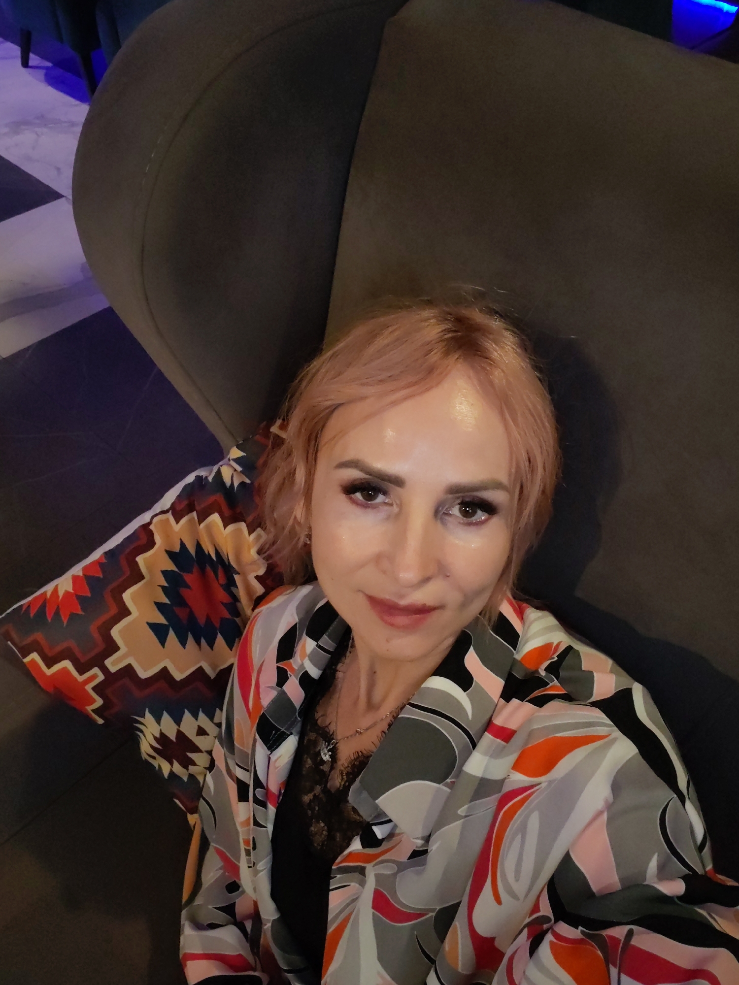 A woman with short, light pink hair and heavy eye makeup is pictured from the chest up, sitting on a dark gray armchair. She is wearing a black lace top and a brightly colored, patterned jacket with abstract shapes in shades of gray, white, pink, orange, and black. To her left is a decorative pillow with a geometric pattern in similar colors. The background shows a dimly lit area with a tiled floor and hints of blue lighting, suggesting an indoor setting such as a lounge or restaurant. The scene is likely in Yoshkar-Ola, Russia, but there is no specific visual information to confirm this location.