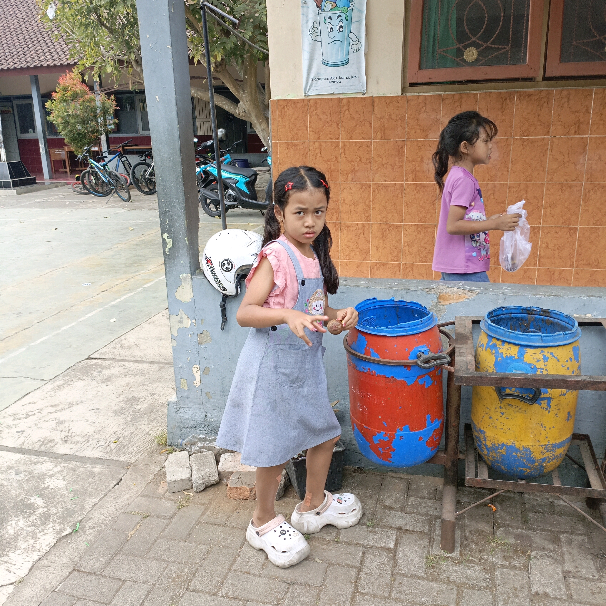 In a courtyard area of what appears to be a school in Salatiga, Indonesia, two young girls are present. The girl in the foreground, on the left, is standing and looking directly at the camera. She has dark hair tied in pigtails with red hair clips. She is wearing a pink t-shirt with short ruffled sleeves and a grey pinafore dress. On her feet are white platform clogs. She is holding a brown object in her right hand. Next to her, to the right, are two large, weathered plastic drums used for waste disposal, one red and blue, the other yellow and blue, placed on a metal stand.
Behind her, to the right and slightly further back, is another girl, also with dark hair pulled back in a ponytail. She is facing to her right, away from the camera. She is wearing a purple t-shirt with a graphic print and appears to be holding a clear plastic bag. She is standing in front of a textured orange tiled wall.
In the background, on the left, bicycles and a motorcycle are parked in front of a building with a red and white facade and a tiled roof. A tree with vibrant red and green foliage is also visible. A white helmet is hanging on a grey pillar in the foreground. The ground is paved with concrete and brickwork, with some scattered debris. A poster depicting a sad trash can is affixed to the building wall above the girls. The scene suggests an outdoor educational or recreational space.