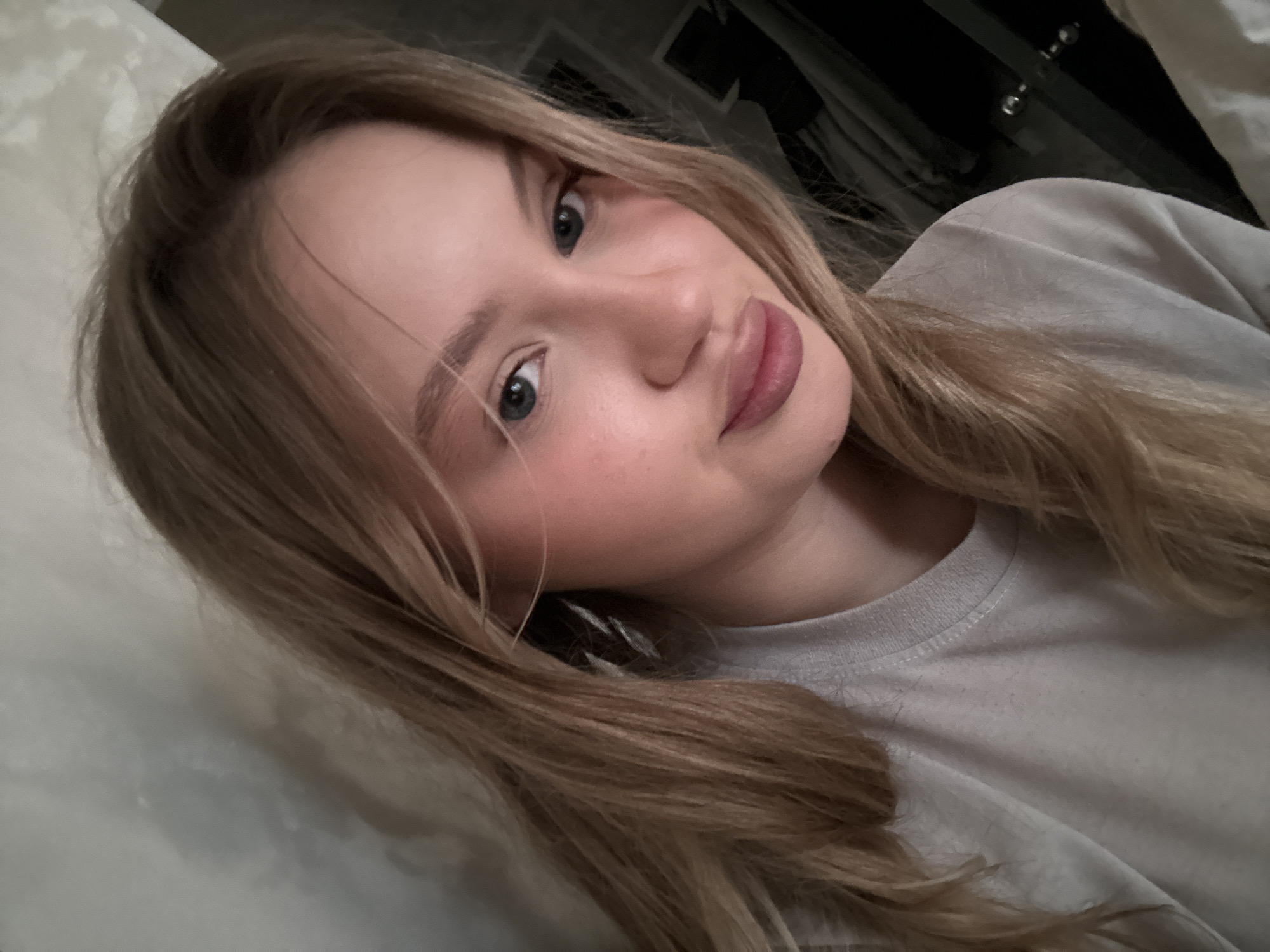 A young woman with blonde hair and blue eyes is taking a selfie indoors. She is wearing a light grey t-shirt. Her hair is down and slightly wavy. Her makeup includes pink blush and a matte pink lipstick. She has a neutral expression. The background is dimly lit and appears to be a room with textured walls and possibly furniture or clothing in the background. The image is cropped to focus on the woman's face and upper chest. There is no indication of the city of Miass, Russia.