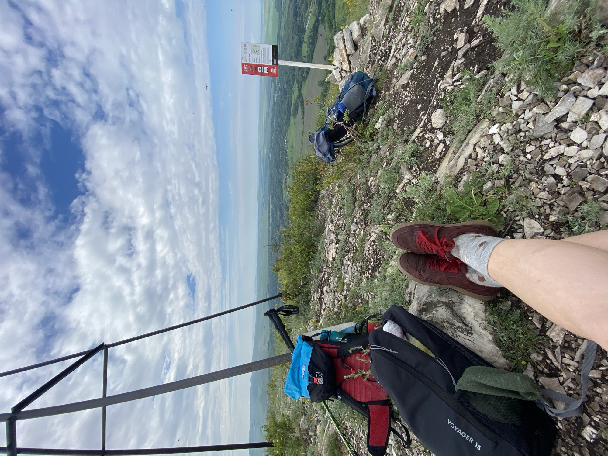 The image depicts a person's legs, clad in maroon sneakers and grey socks, resting on a rocky, grassy slope. In the foreground, to the left, is a black backpack with the label "VOYAGER 15". Behind this backpack, a red and black hiking backpack is visible, topped with a bright blue item. Trekking poles are propped up against a metal structure on the left side of the frame. Further to the right, another backpack, predominantly grey, leans against a bush near a signpost. The signpost displays text and graphics, indicating "5УТ" and "Гора Хос". In the background, a vast, green valley stretches out under a partly cloudy sky, with distant hills visible on the horizon. The overall scene suggests a hiking or mountaineering excursion in a natural, elevated environment.