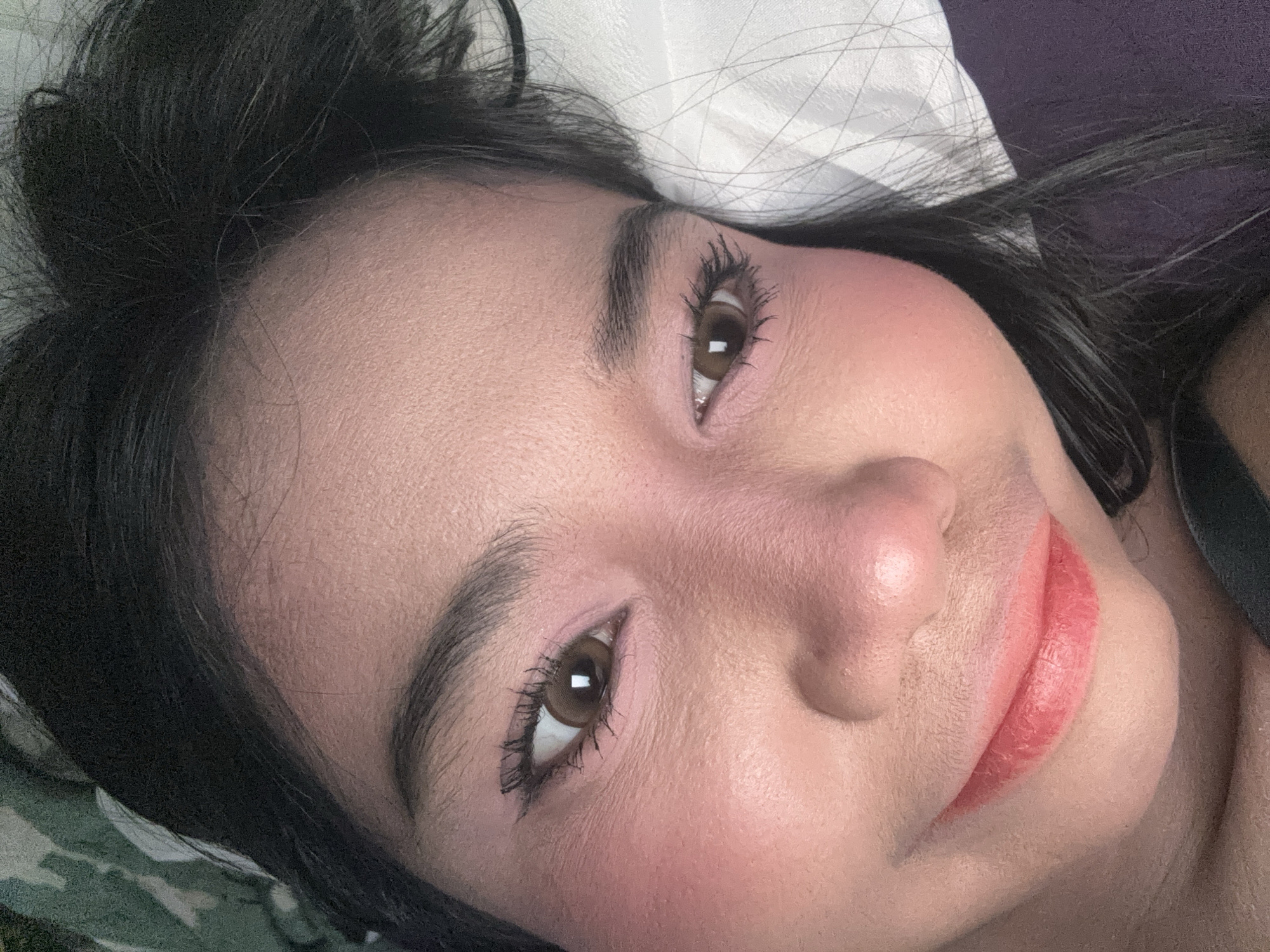 A close-up, overhead perspective captures the face of an individual, likely female, resting in an indoor setting within North Jakarta, Indonesia. The subject is seen lying down with a slight head tilt, looking directly towards the camera with a relaxed expression and a subtle smile. Both eyes are open, featuring dark brown irises with visible pupils. The eyelashes are dark and appear volumized by mascara, complemented by a precise winged eyeliner. The eyebrows are dark, full, and well-defined. The lips are full, adorned with a reddish-pink lipstick, and are slightly parted. The skin exhibits a fair to medium tone with visible texture, and a prominent pink blush is applied to the cheeks and across the bridge of the nose. Dark, straight hair is visible flowing away from the face on the left and bottom right sides of the frame. The immediate environment includes a dark, possibly purple, fabric on the lower left, a white fabric with subtle linear patterns on the bottom and right, and a dark green camouflage-patterned fabric on the upper right, suggesting textiles like clothing or bedding.